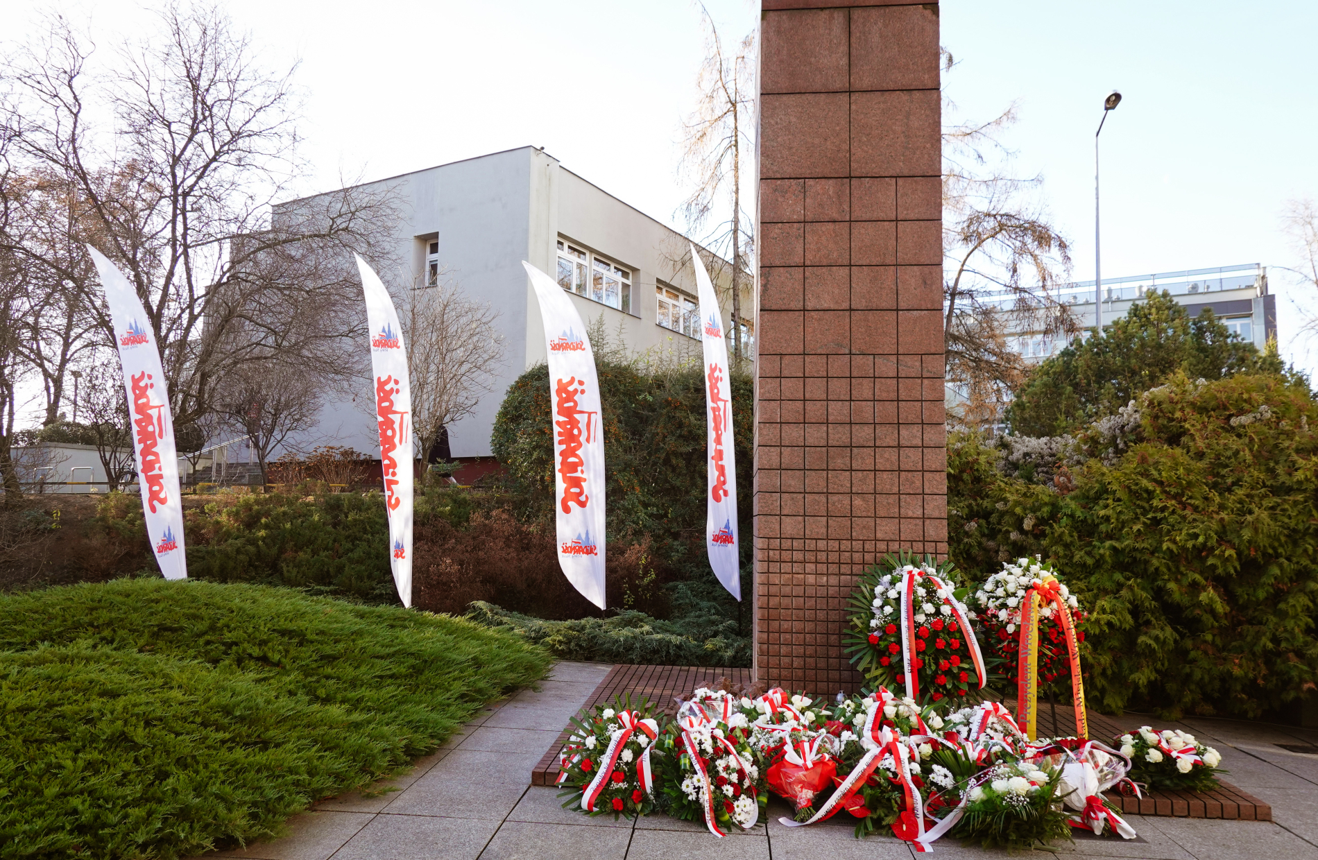 Zdjęcie pomnika ze złożonymi kwiatami, w tle flagi Solidarności
