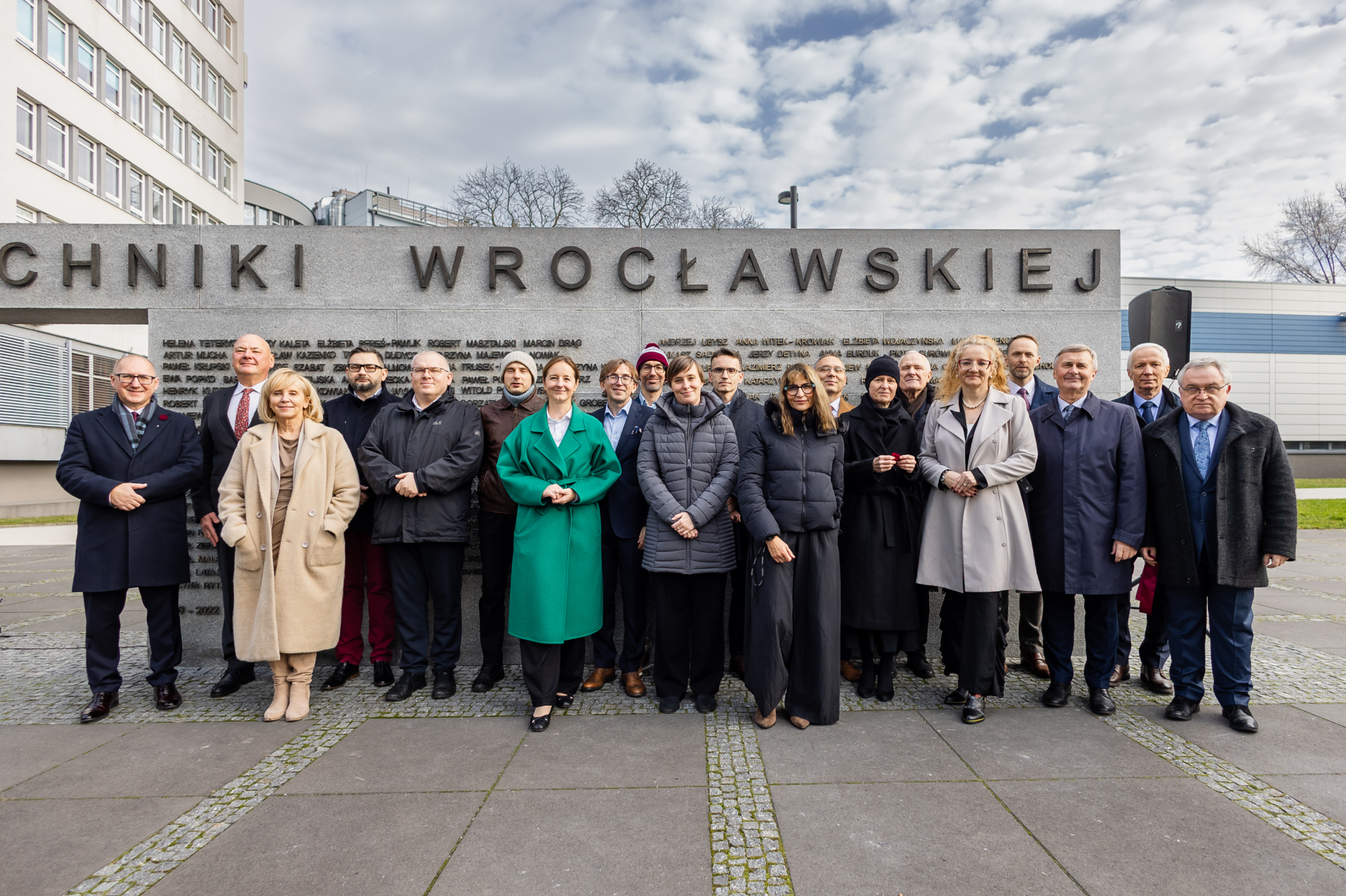 Grupa osób, kobiet i mężczyzn, stoi razem do pamiątkowego zdjęcia przed dużym kamiennym murem z wyrytymi napisami.