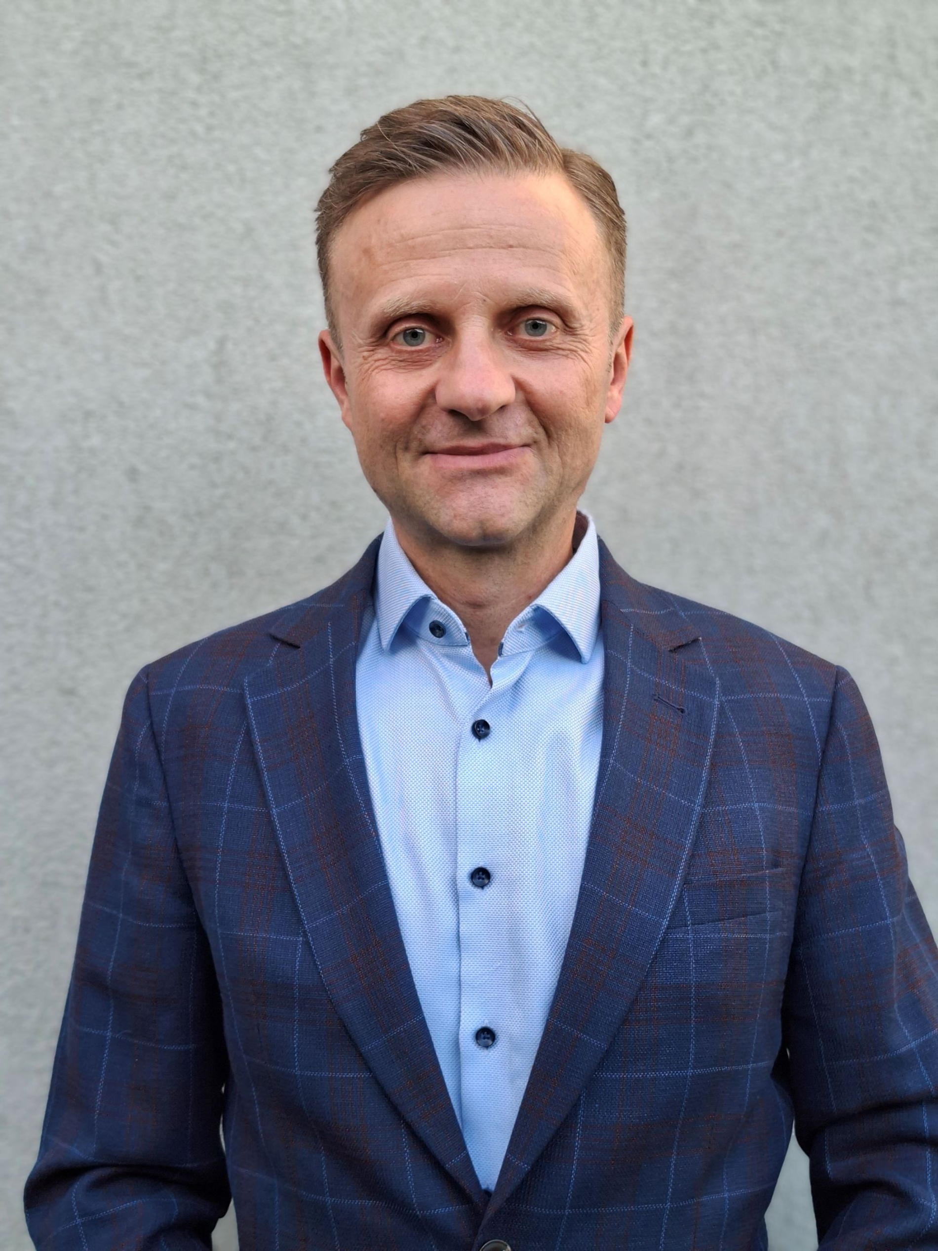 Mężczyzna w granatowej marynarce w kratę i błękitnej koszuli, uśmiecha się lekko do obiektywu na tle szarej ściany.