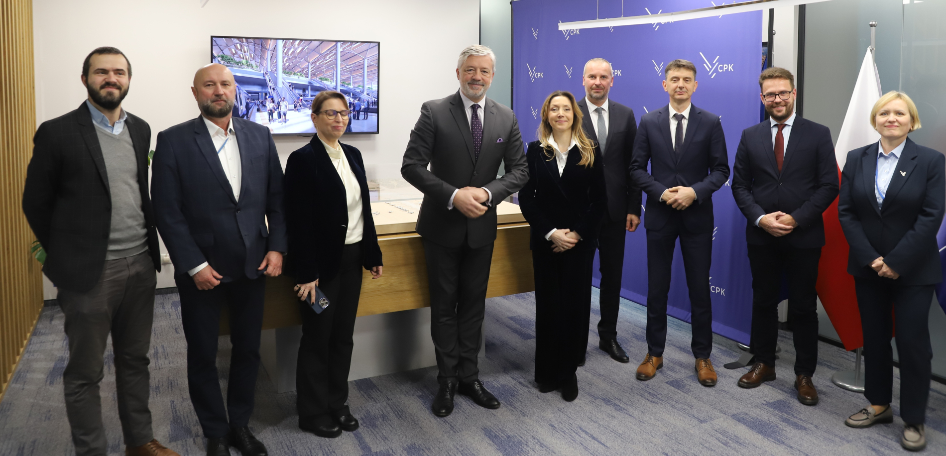 Dziewieć osób w strojach biznesowych stoi na spotkaniu w nowoczesnym biurze CPK, z logo firmy na ścianie