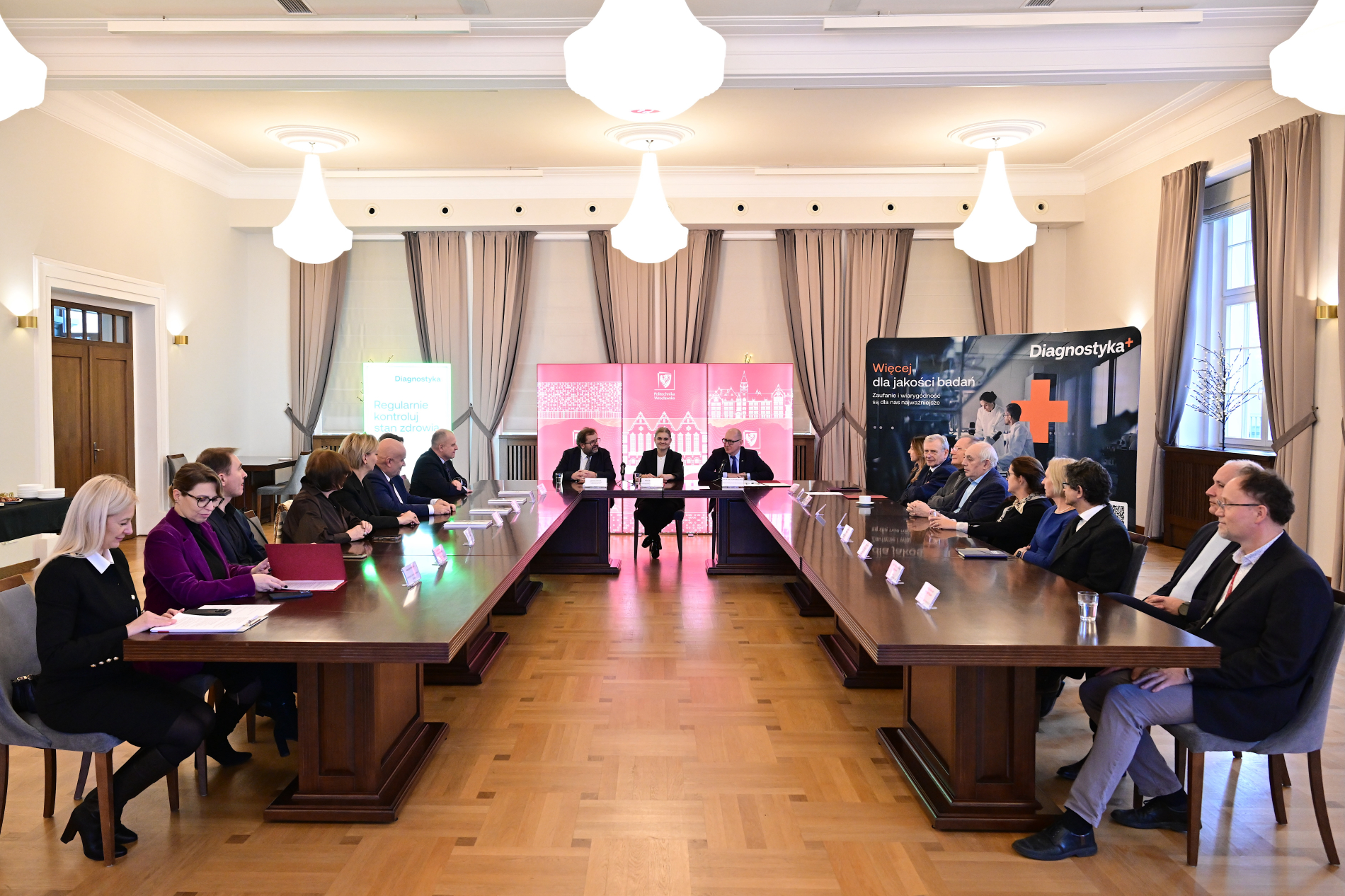 Dwie grupy osób siedzą przy długim, połączonym stole naprzeciwko siebie. W tle widoczne banery promocyjne.