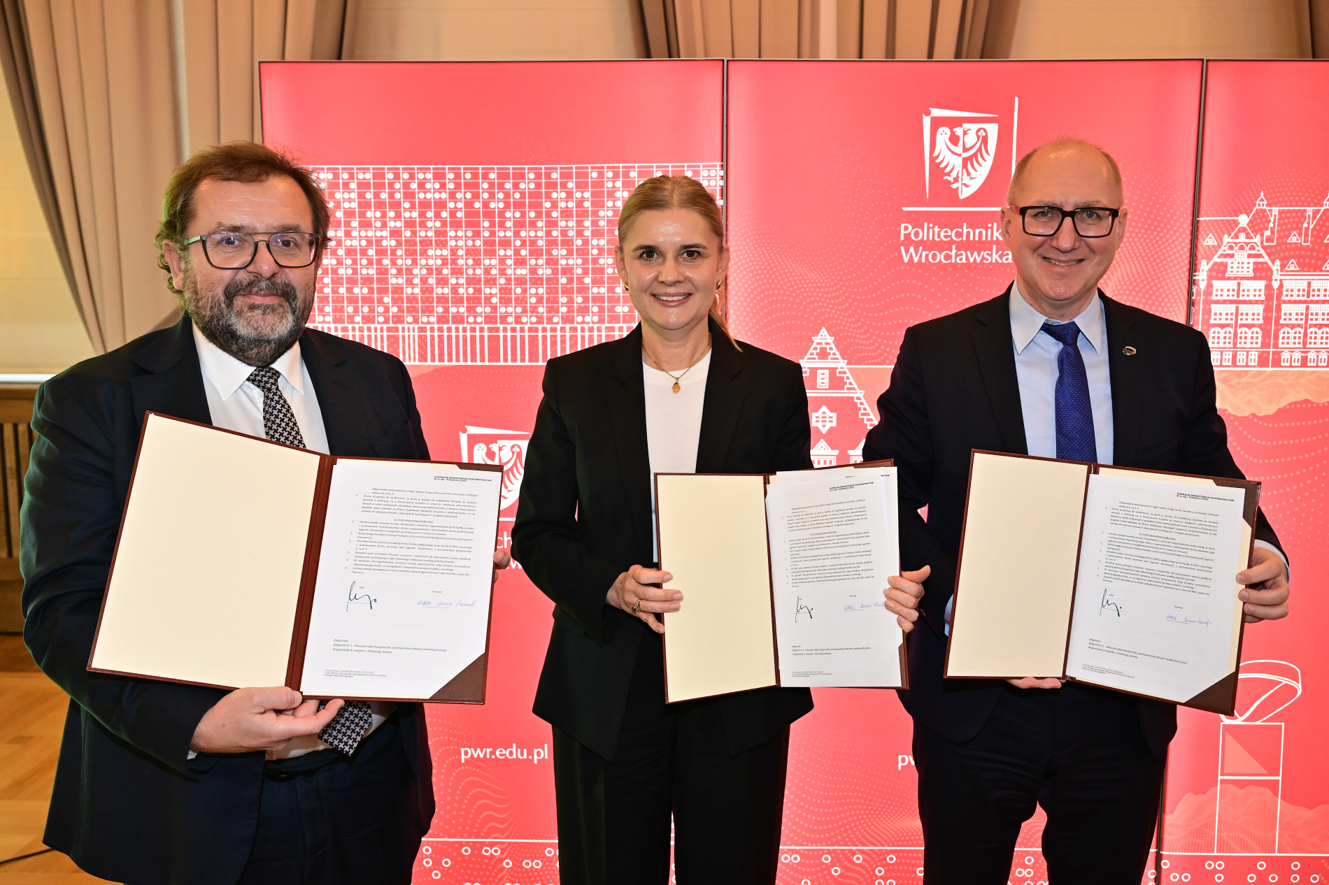 Trzy osoby (dwóch mężczyzn i kobieta w środku) w formalnym stroju trzymają otwarte dokumenty, prawdopodobnie po podpisaniu umowy. W tle logo Politechniki Wrocławskiej.