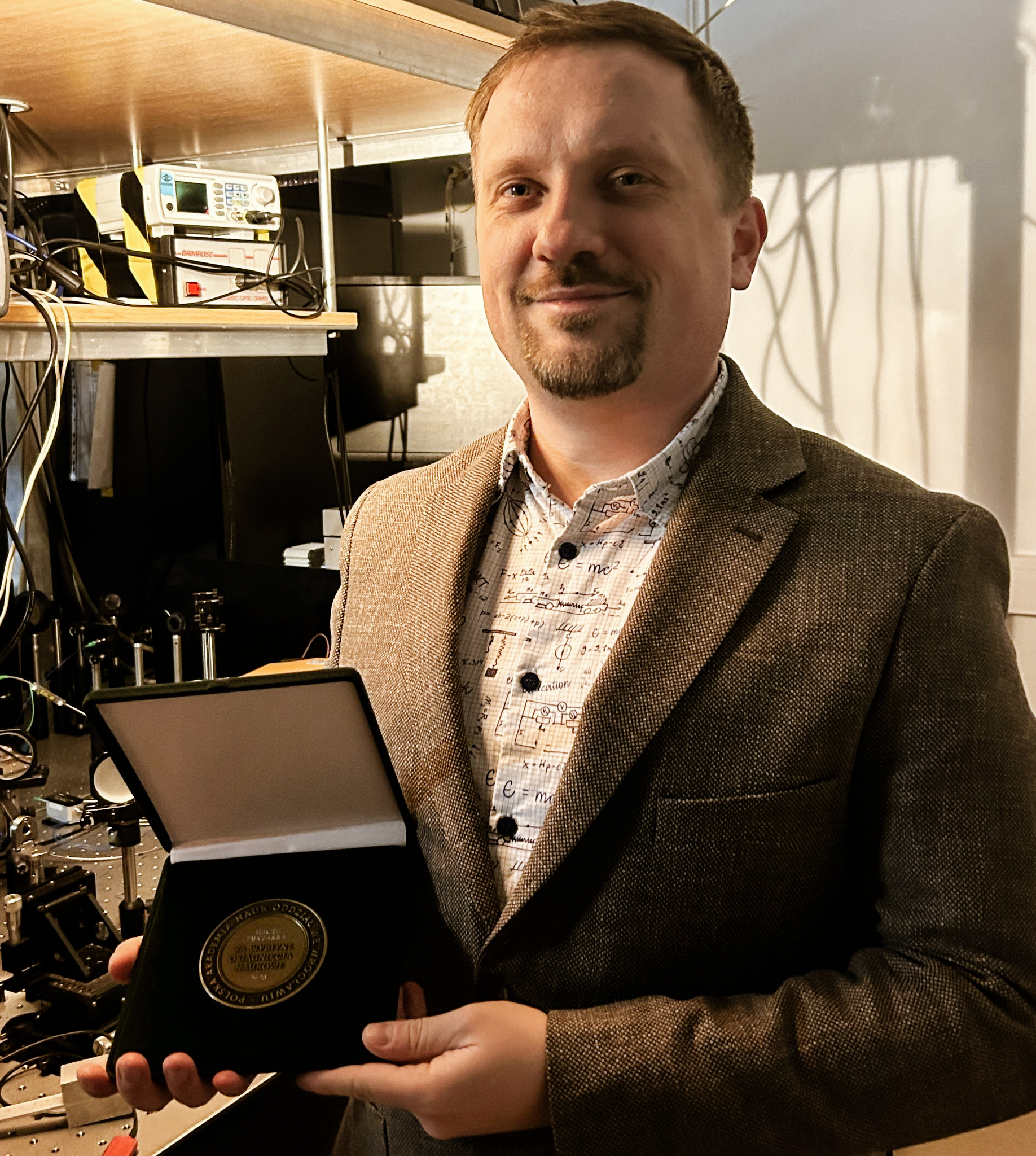 Mężczyzna w marynarce prezentuję medal, w tle widać sprzęt badawczy.