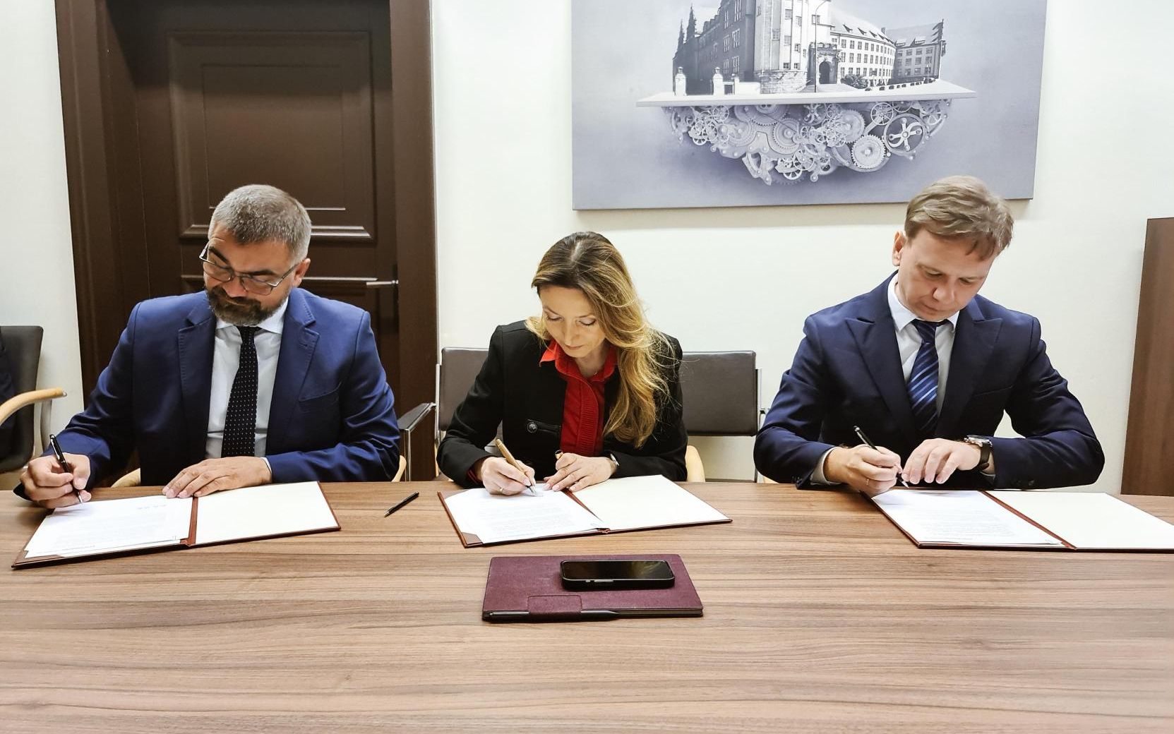 Trzy osoby w strojach biznesowych siedzą przy drewnianym stole i składają podpisy na dokumentach.