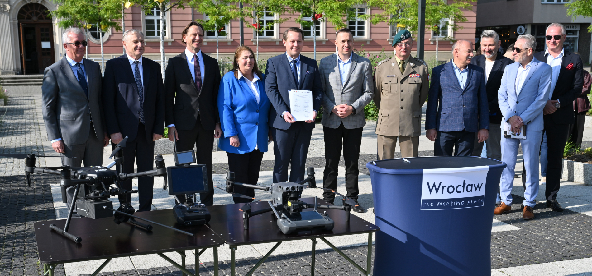 Zdjecie przedstawia grupę mężczyzn i jedną kobietę stojących za stołem z dwoma dronami i ekranami sterującymi, na tle budynku.