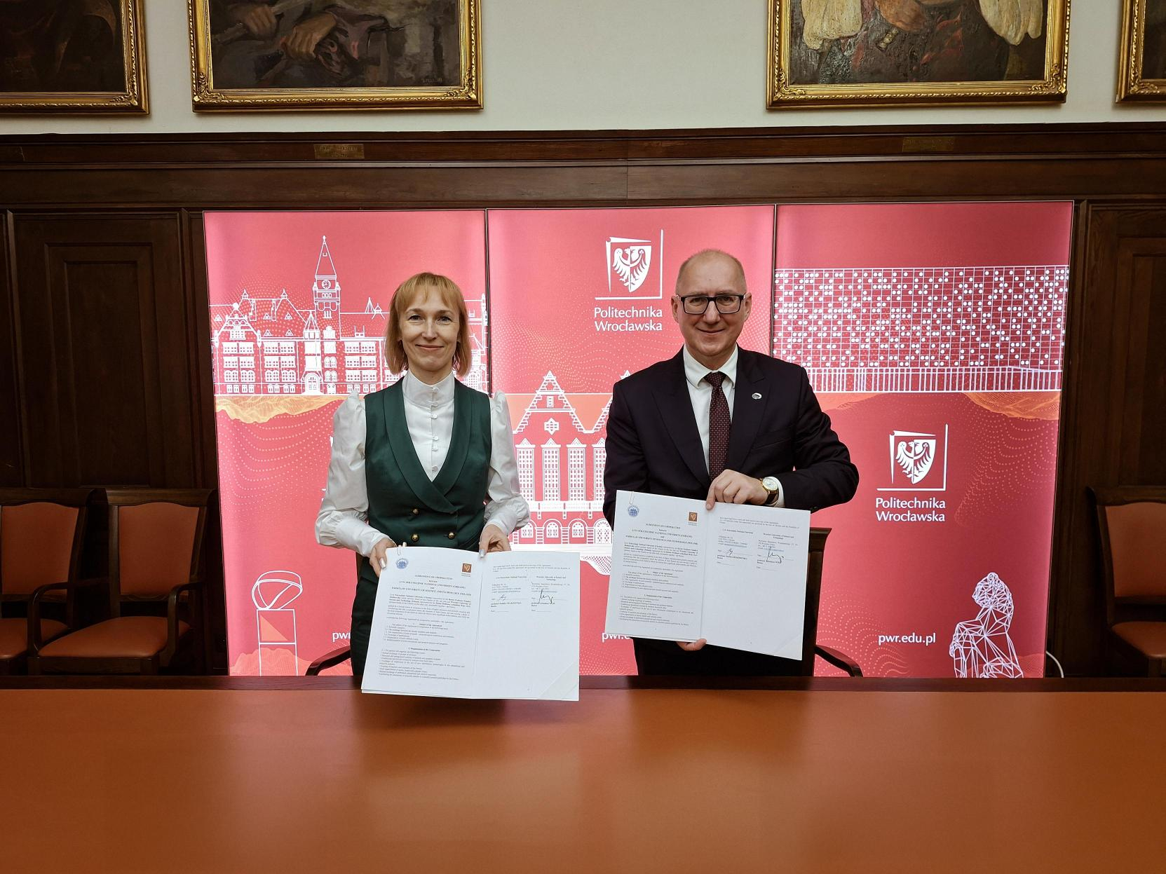 Dwie osoby, kobieta i mężczyzna, stoją za stołem i trzymają podpisane dokumenty. W tle widoczne logo Politechniki Wrocławskiej.