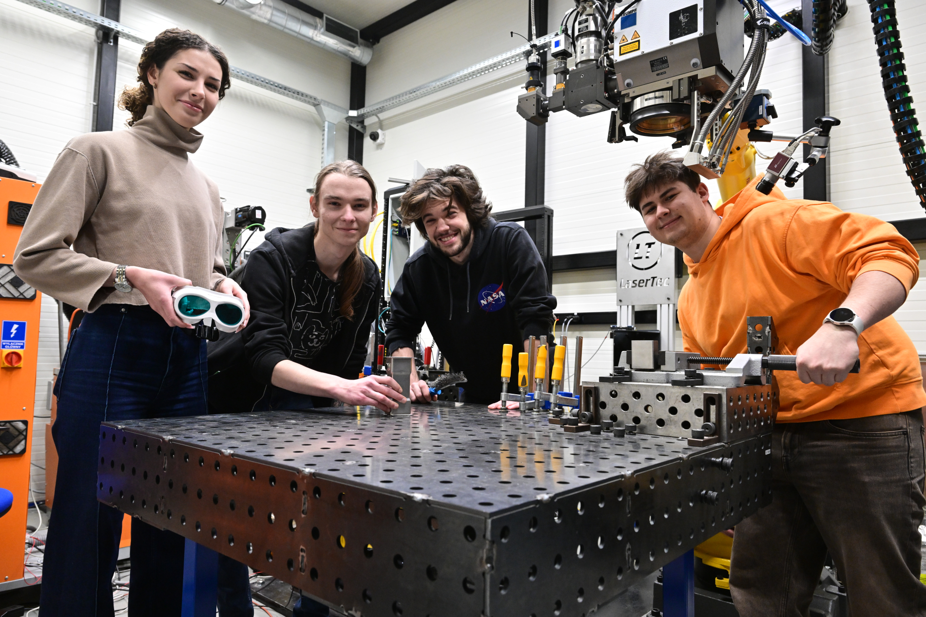 Czworo uśmiechniętych młodych ludzi pozuje w nowoczesnym laboratorium inżynieryjnym przy perforowanym stole warsztatowym.