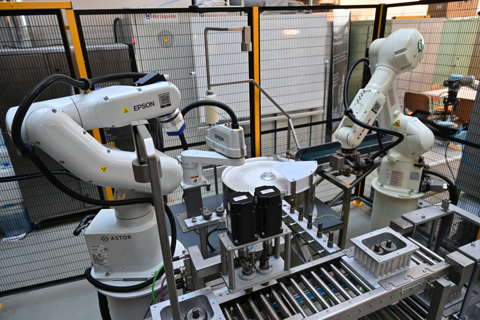 Trzy białe ramiona robotów przemysłowych pracujące na zrobotyzowanym stanowisku dydaktycznym.