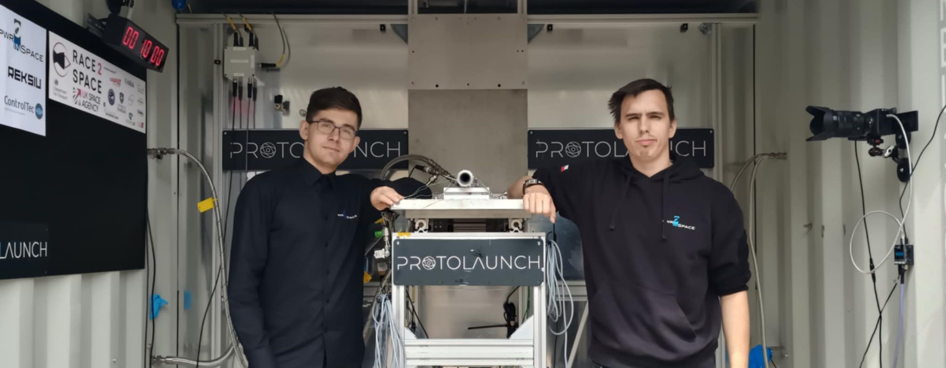 Dwóch młodych mężczyzn stoi obok testowanego silnika rakietowego w laboratorium. Na ścianie w tle widoczne są logotypy PWR in Space i Reksio. Mężczyźni są uśmiechnięci i opierają się o sprzęt, z widocznym dumnym wyrazem twarzy.