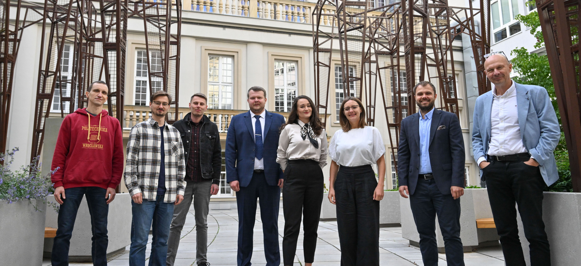 Osiem osób stoi obok siebie na dziedzińcu. W tle widoczny jasny, zabytkowy budynek z oknami i nowoczesna, metalowa konstrukcja. Uśmiechnięta grupa w strojach biznesowych i casualowych.