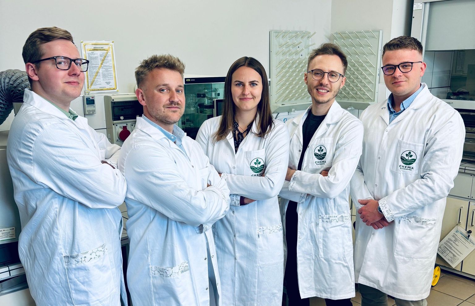 Grupa studentów, ubrana w fartuchy laboratoryjne z zielonym logo "CHEMIA", pozuje z założonymi rękami w dobrze oświetlonym laboratorium.