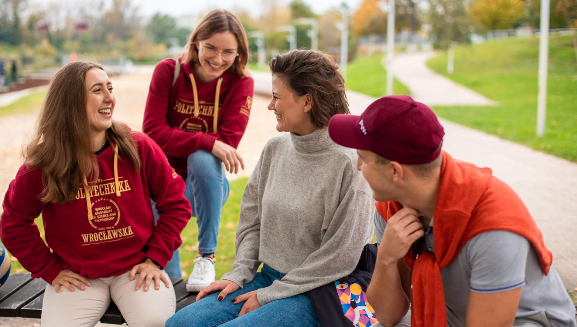Studenci w bluzach z logo Politechniki Wrocławskiej i w swobodnym stroju siedzą razem na ławce.
