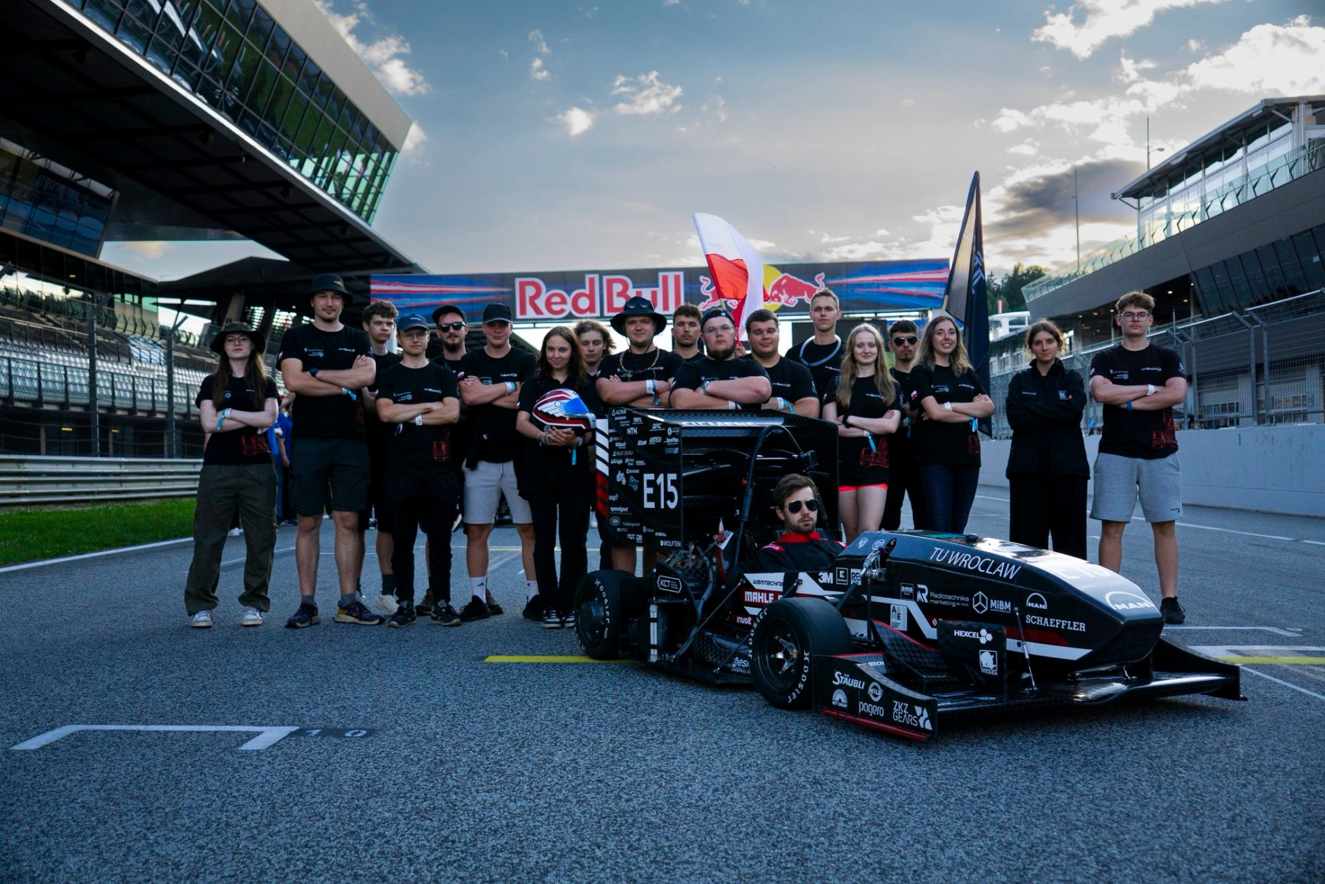 Duża grupa studentów soi na torze wyścigowym, a przed nimi znajduje sie mały bolid wyścigowy z kierowcą.