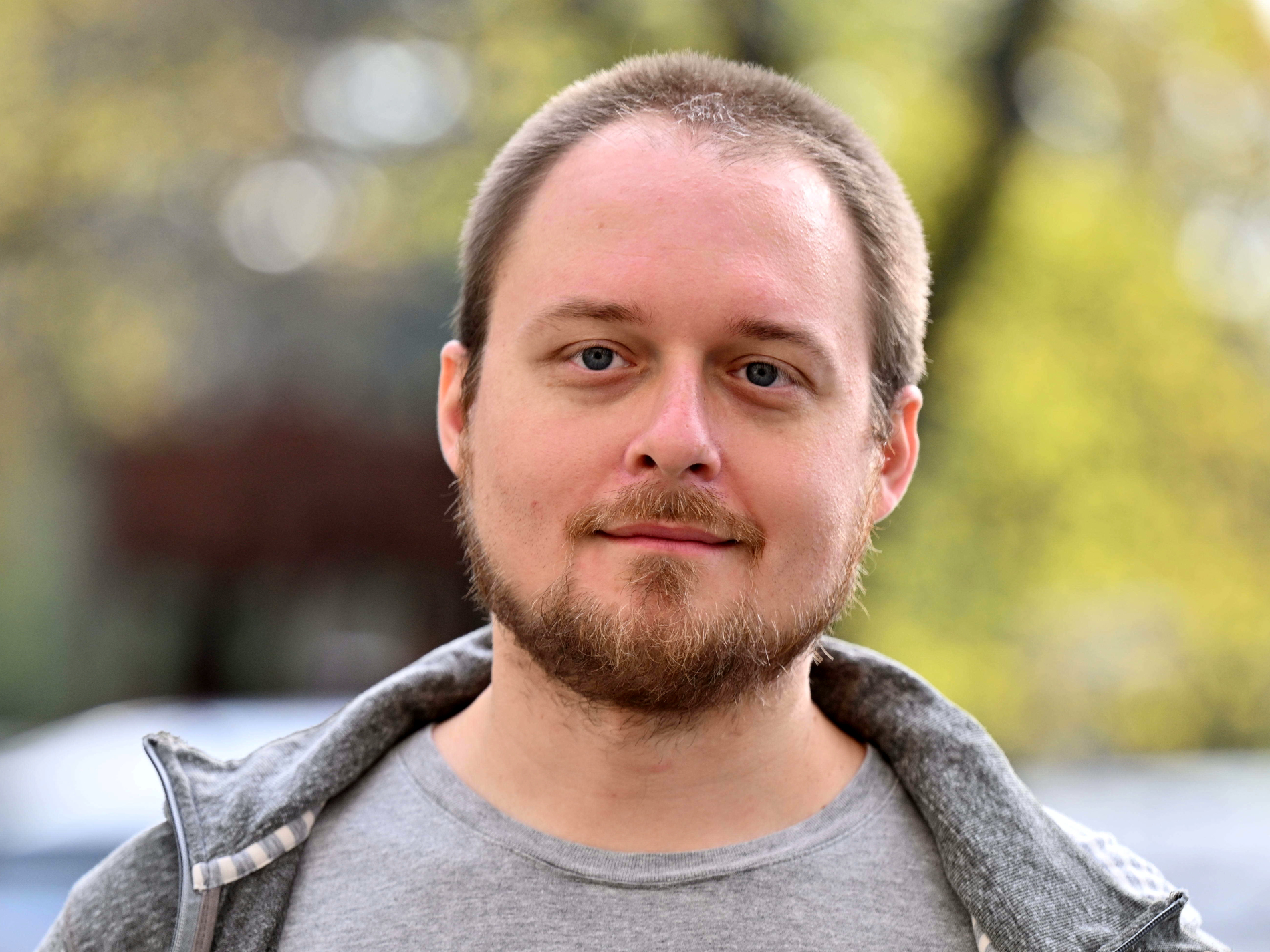 Mężczyzna o jasnej karnacji, krótkich, ciemnych włosach i brodzie, ubrany w szarą bluzę z kapturem, patrzy prosto w obiektyw. Tło jest rozmyte, z widocznymi jasnymi punktami.