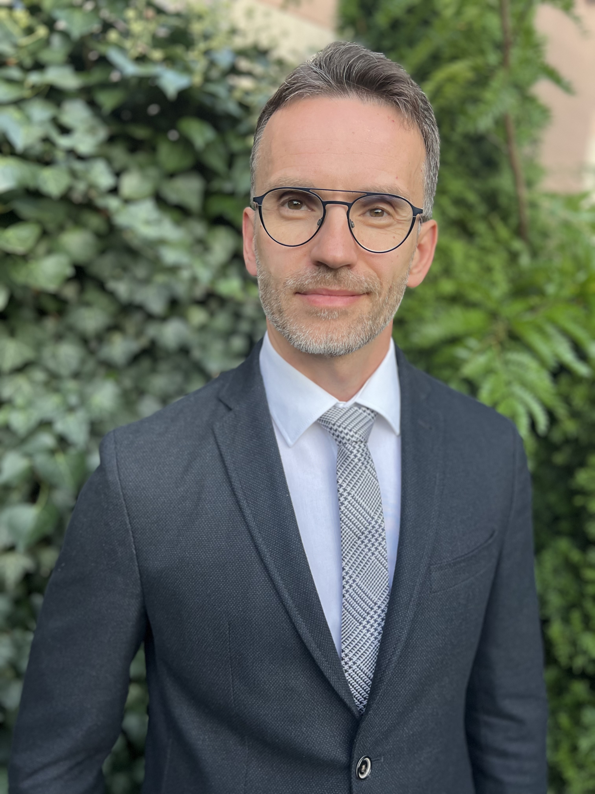 Portret uśmiechniętego mężczyzny w okularach i eleganckim garniturze. Stoi na tle ściany porośniętej zielonym bluszczem.