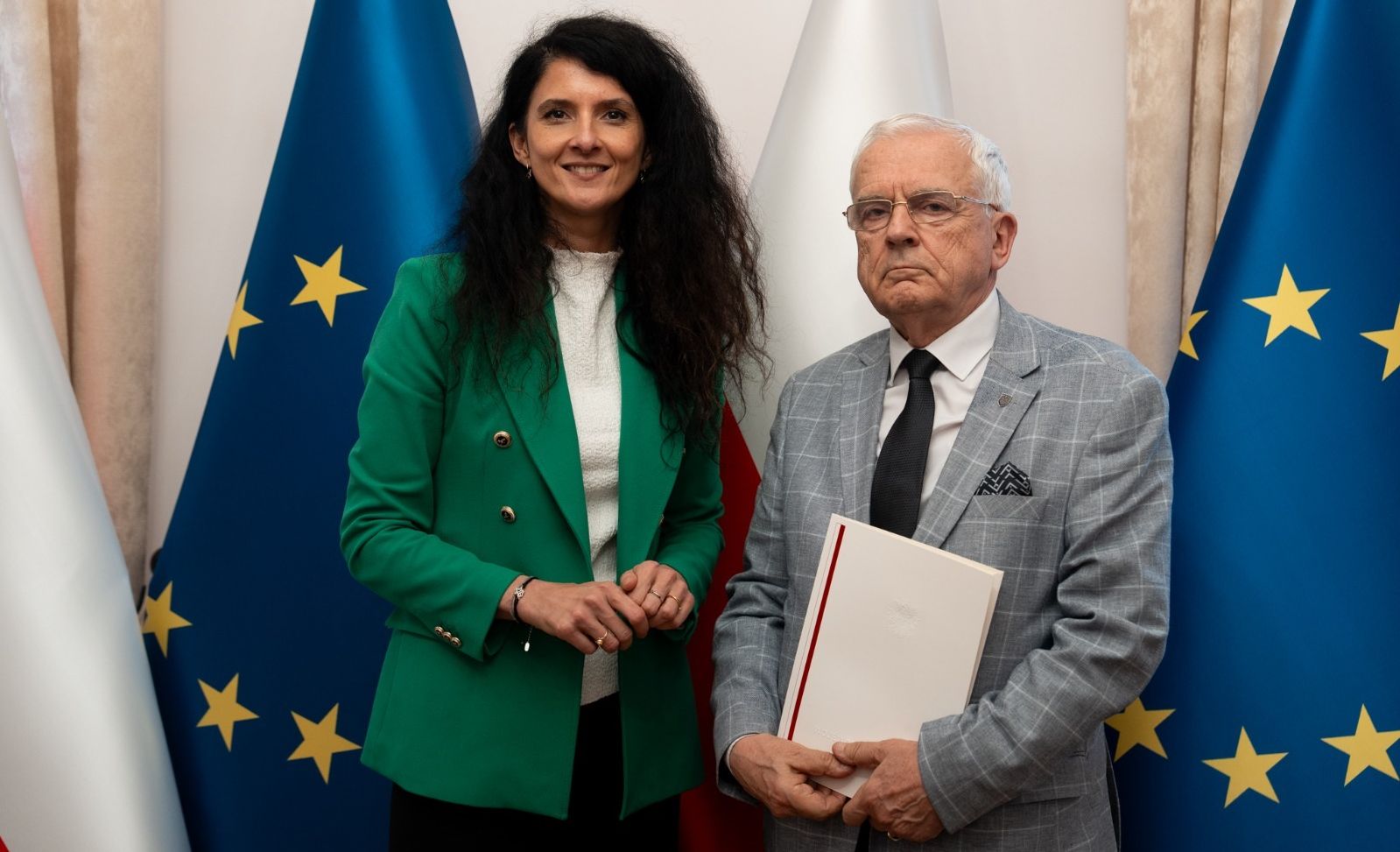 Elegancko ubrana kobieta w zielonej marynarce stoi obok starszego mężczyzny w szarej marynarce, który trzyma teczkę. W tle widoczne są flagi Polski i Unii Europejskiej. Oboje patrzą w kierunku aparatu.