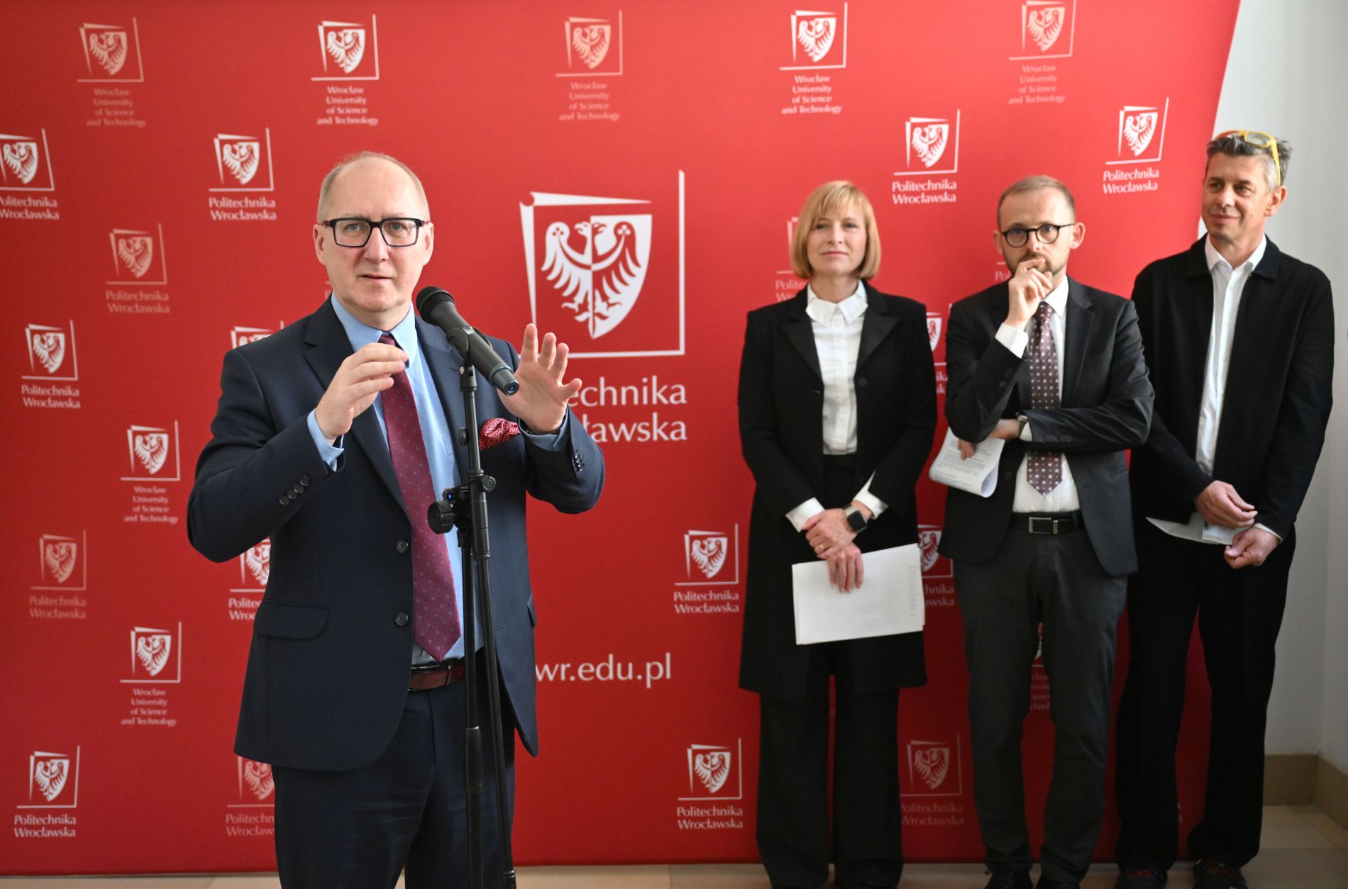 Zdjęcie rektora przemawiającego w holu na tle czerwonej ścianki uczelni