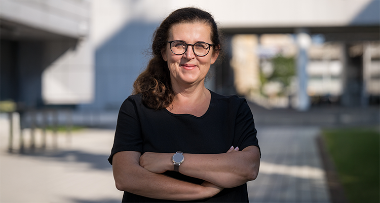 Kobieta w okularach, z uśmiechem i skrzyżowanymi rękami. Ma na sobie czarną koszulkę. Stoi na zewnątrz, w tle widać nowoczesny budynek.