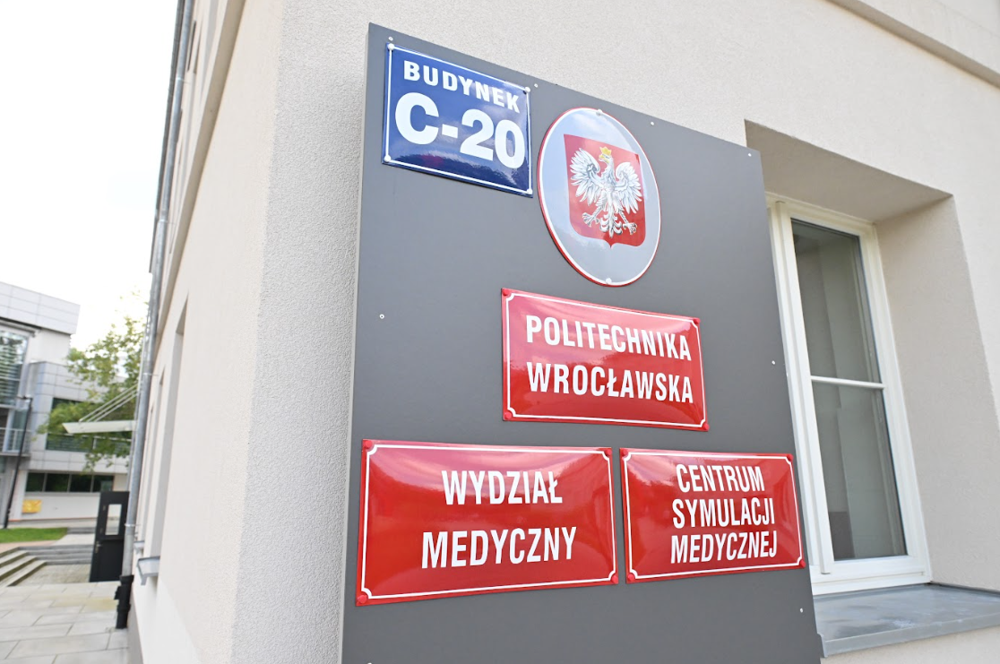 Tablica informacyjna na budynku Politechniki Wrocławskiej. Widoczne są tabliczki z napisami: „Politechnika Wrocławska”, „Wydział Medyczny” oraz „Centrum Symulacji Medycznej”. Na górze znajduje się numer budynku, C-20, oraz godło Polski.
