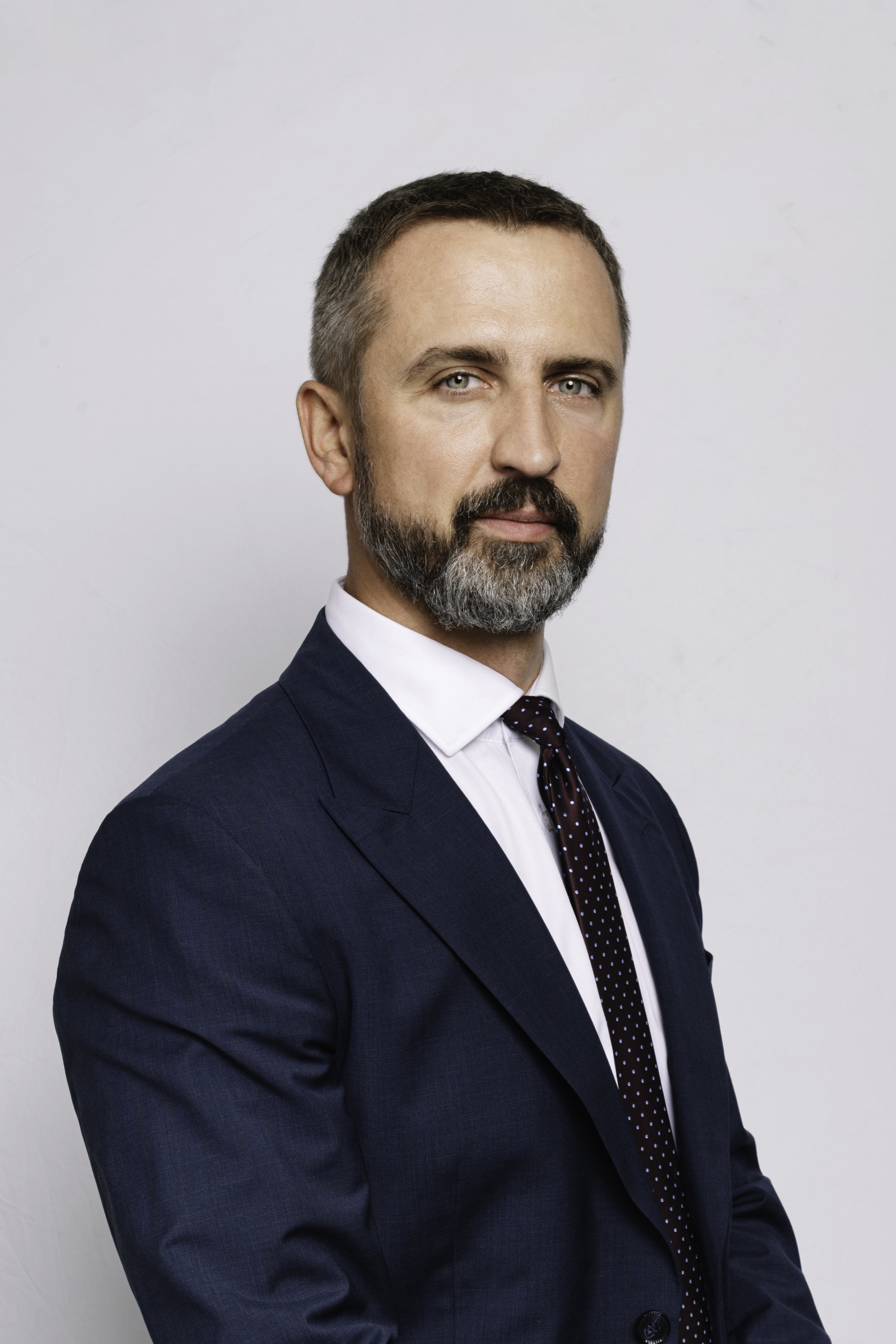 Mężczyzna w średnim wieku, uśmiechnięty, w granatowej marynarce na białej koszuli, pozujący na jasnym tle.
