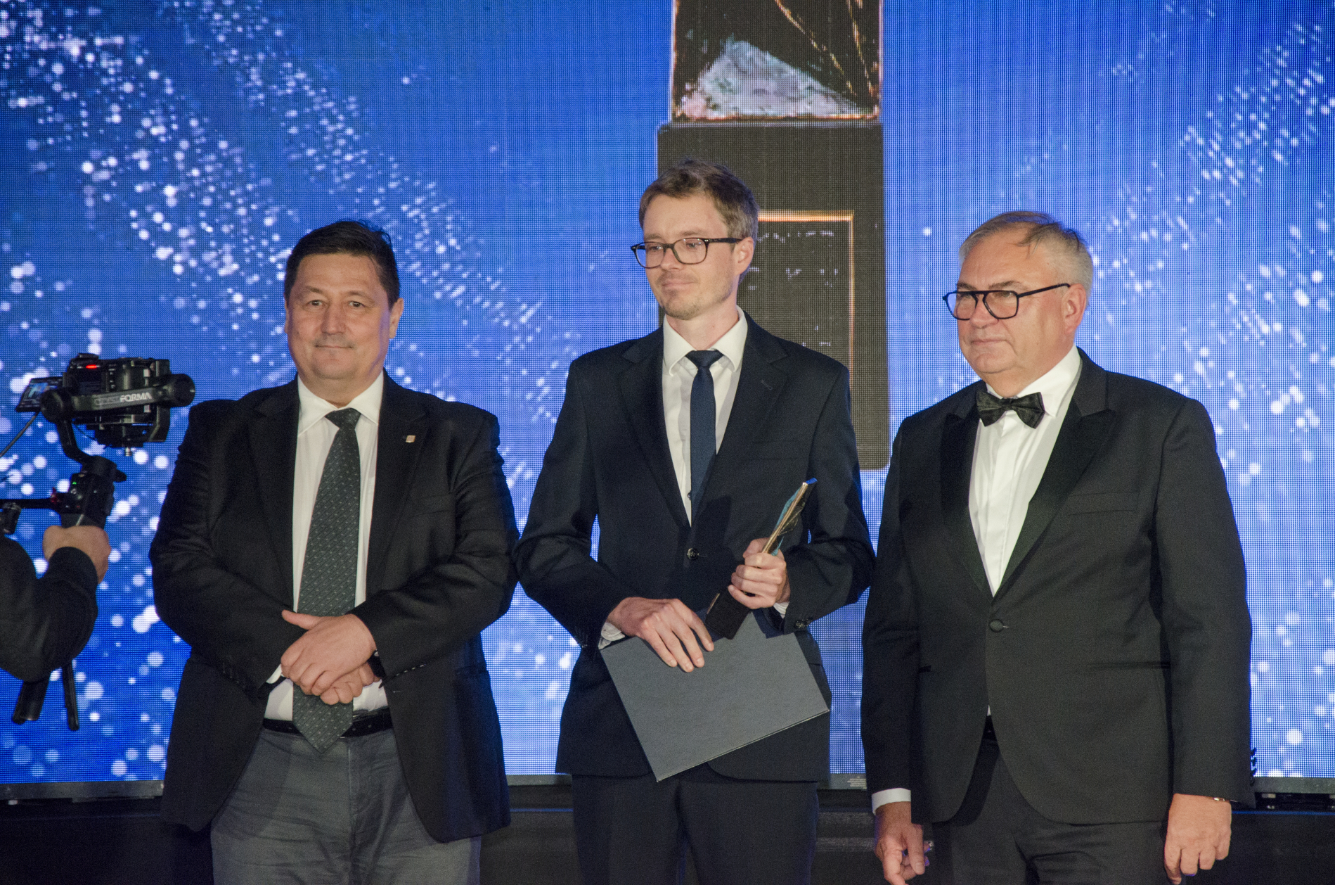 Trzech mężczyzn w strojach wieczorowych stoi na scenie na tle świecącego, niebieskiego ekranu. Środkowy mężczyzna trzyma nagrodę i dyplom, prawdopodobnie podczas ceremonii wręczenia.