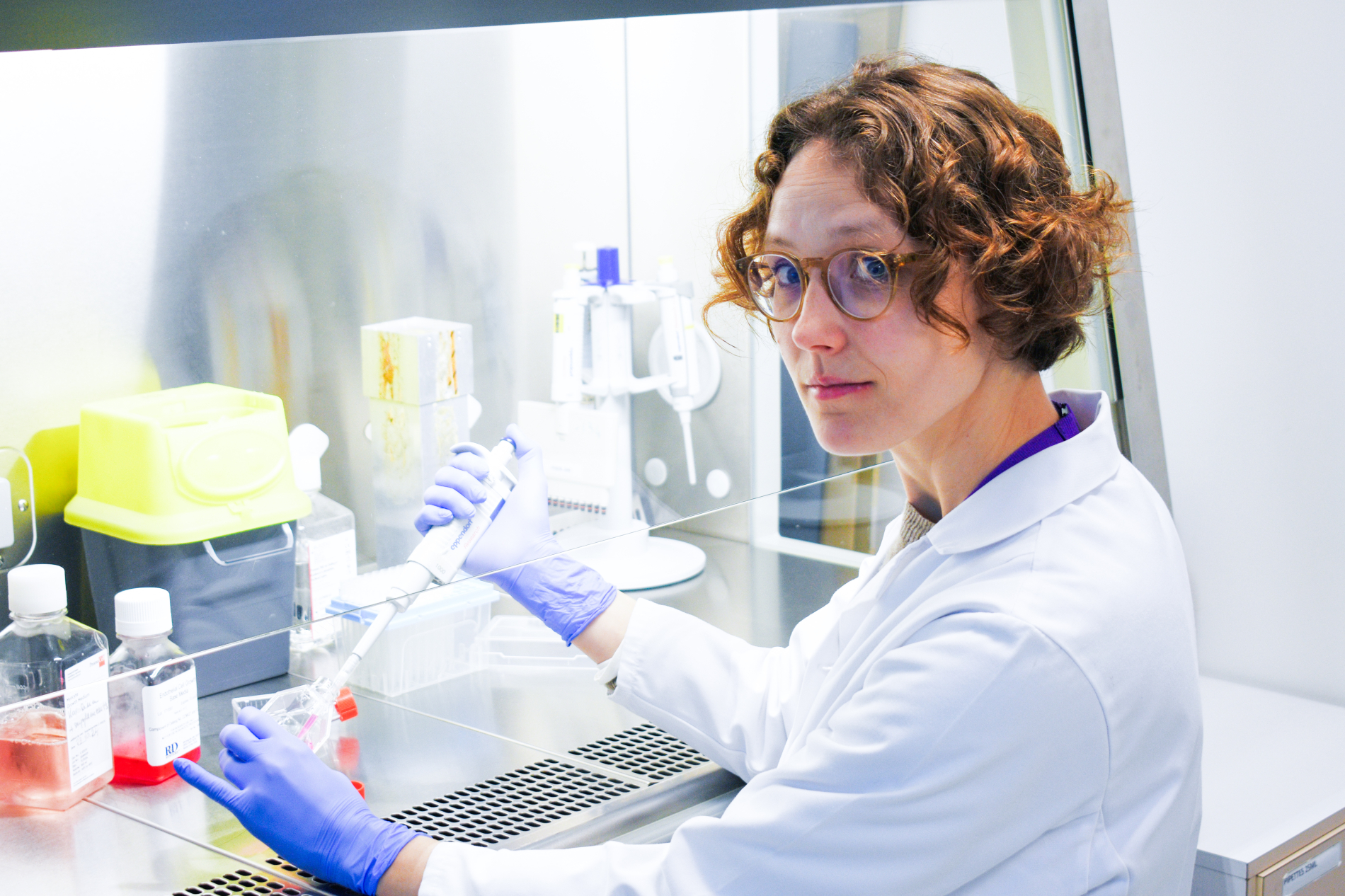 Kobieta w okularach i białym fartuchu laboratoryjnym pipetuje ciecz w komorze laminarnej.