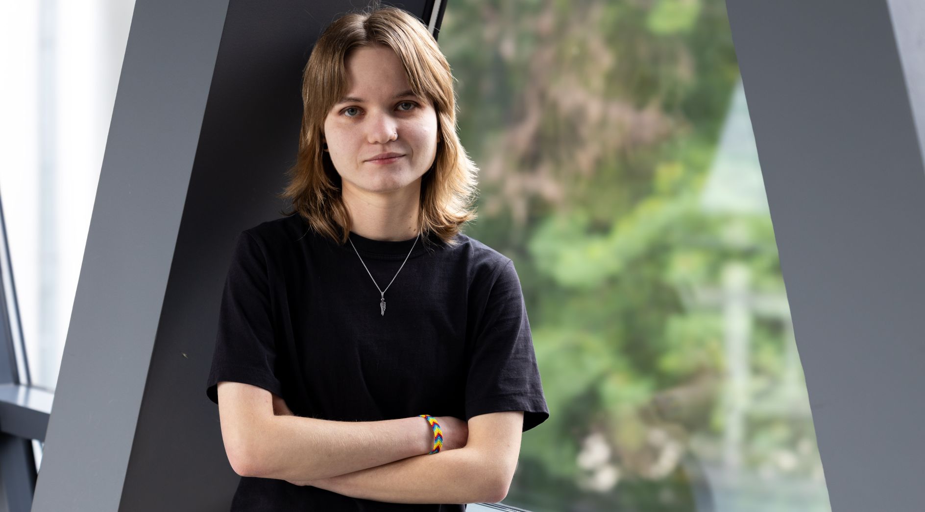 Młoda kobieta z jasnobrązowymi włosami i zawieszonym na szyi wisiorkiem w kształcie klucza, ubrana w czarny T-shirt, stoi, opierając się o ciemną ścianę obok dużego okna. Ma skrzyżowane ramiona. W tle widać zamazane drzewa i zieleń za oknem.