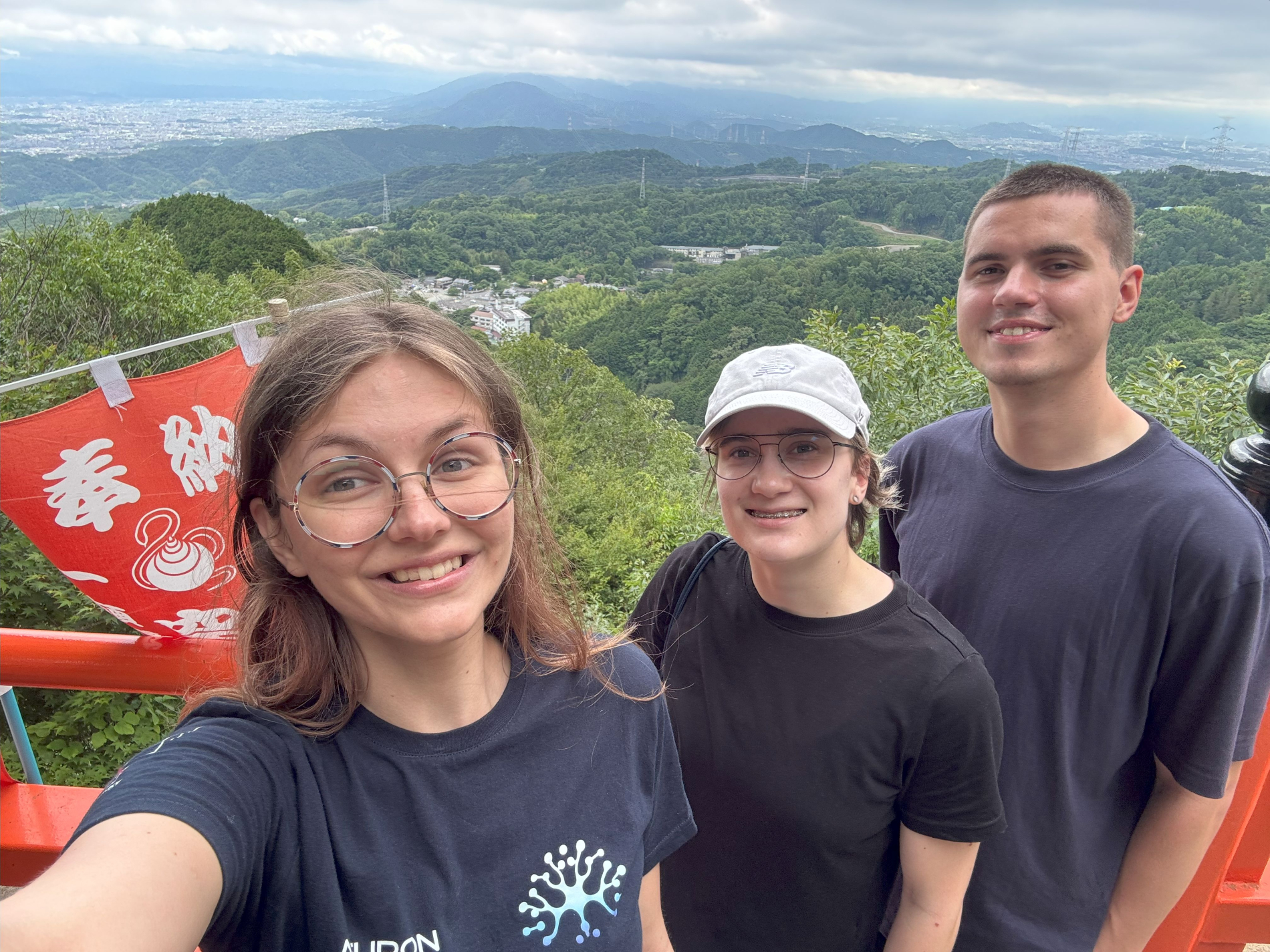 Trzy młode osoby uśmiechają się do aparatu, robiąc sobie selfie na tle górzystego, zielonego krajobrazu. Po lewej stronie widoczny jest fragment czerwonej konstrukcji z japońskimi napisami.