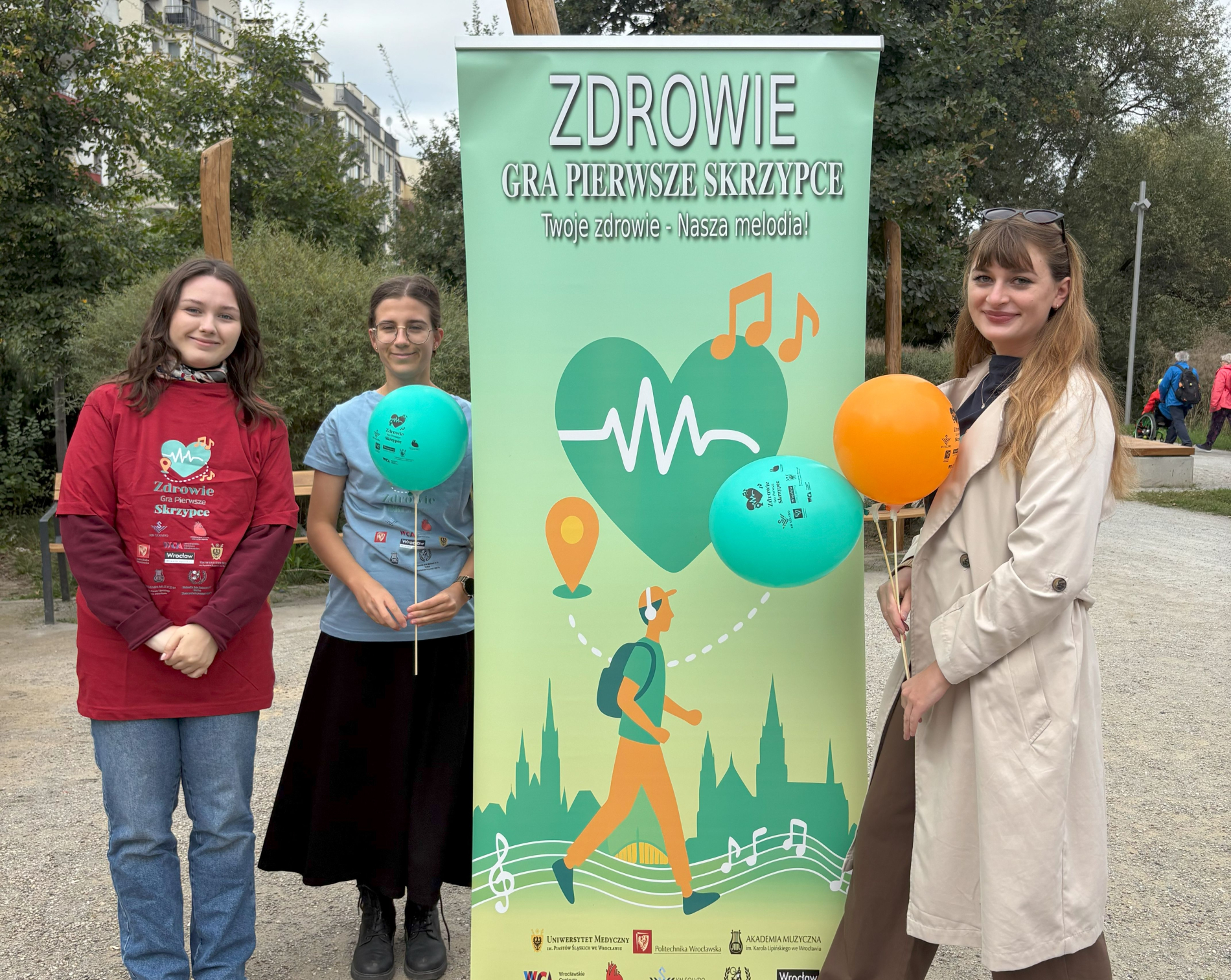 Trzy młode kobiety stoją przy dużym zielonym bannerze z hasłem "Zdrowie gra pierwsze skrzypce". Dwie z nich trzymają turkusowe i pomarańczowe balony. 