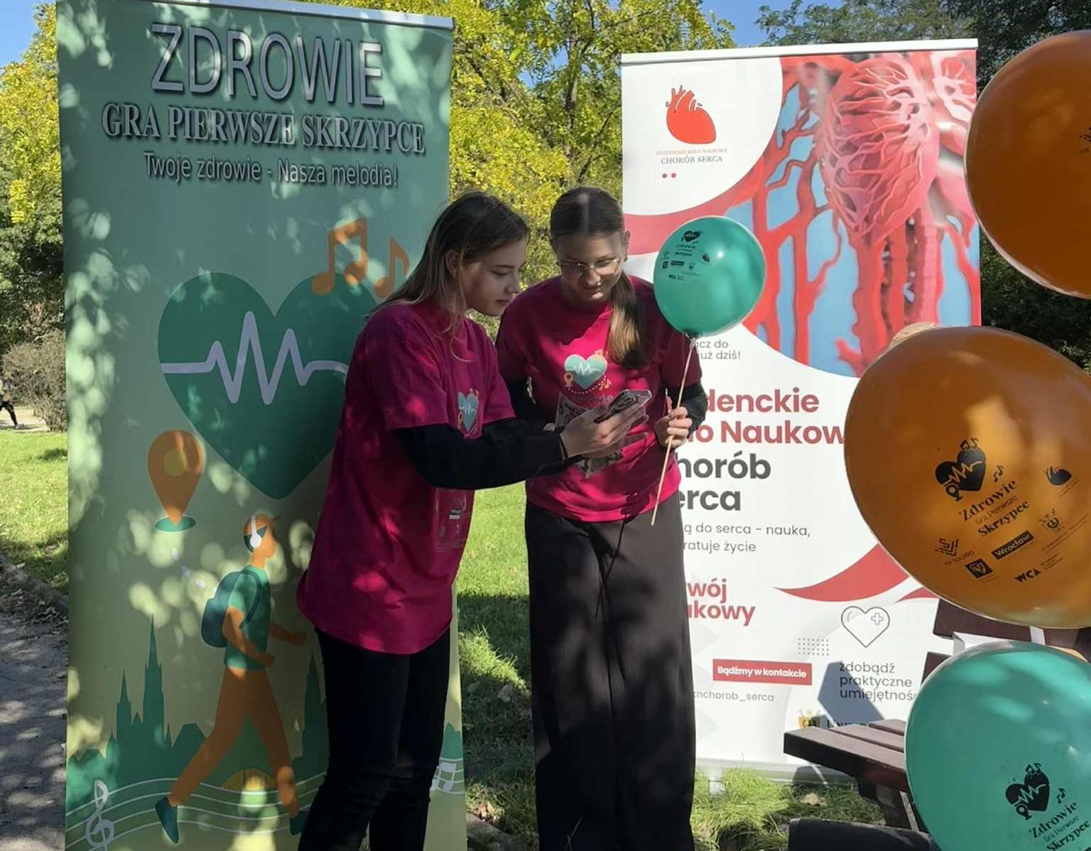 Dwie kobiety w różowych koszulkach sprawdzają coś na smartfonie. Stoją w parku między dwoma dużymi bannerami promocyjnymi, z których jeden ma hasło "Zdrowie gra pierwsze skrzypce".