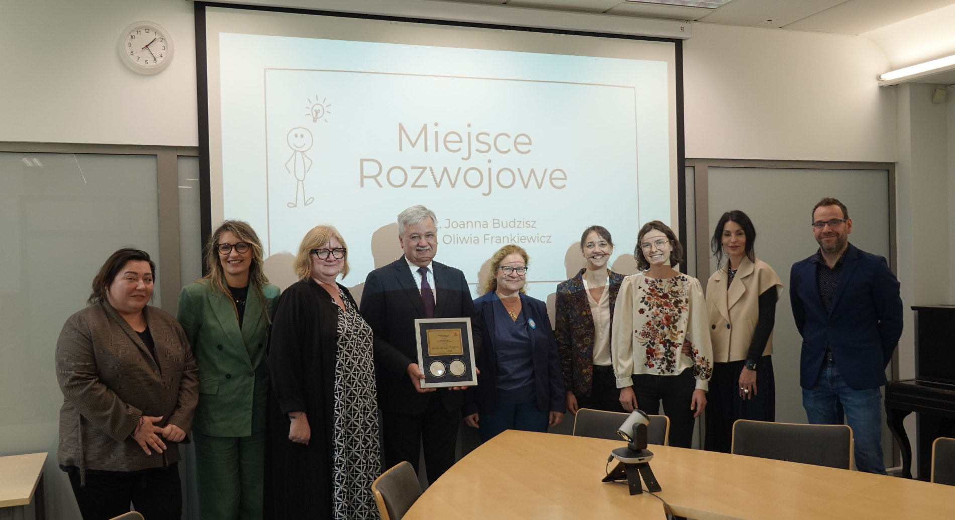 Wręczenie nagrody "Miejsce Rozwojowe". Grupa dziewięciu osób, w tym mężczyzna trzymający tabliczkę z nagrodą i dwa medale, stoi w sali konferencyjnej. 