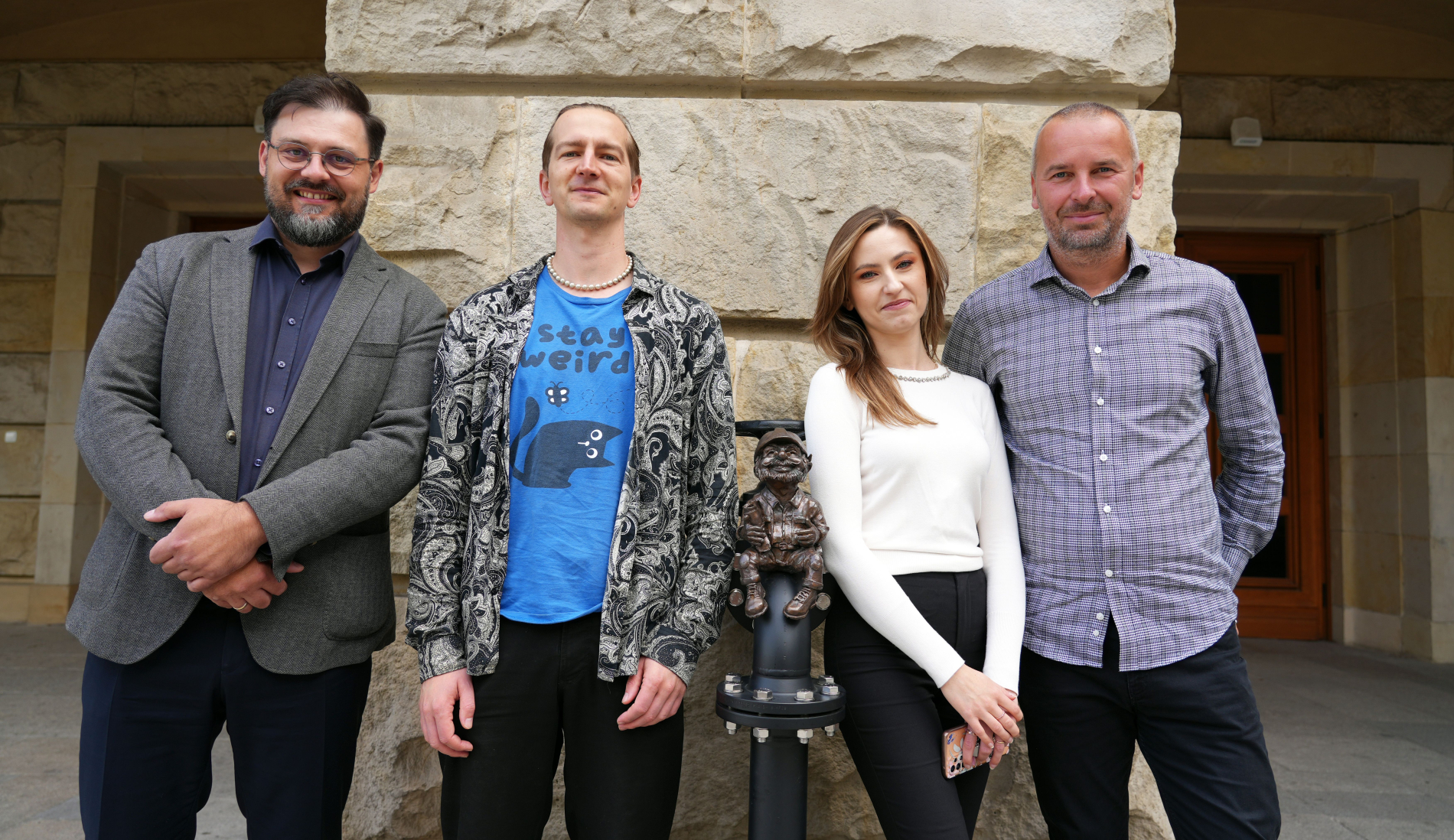 Na zdjęciu widoczna jest grupa czterech osób: trzech mężczyzn i jednej kobiety, pozujących na tle kamiennej ściany. W centrum zdjęcia, między dwiema osobami, znajduje się rzeźba małego, siedzącego gnoma. Wszyscy uśmiechają się, patrząc w stronę obiektywu.