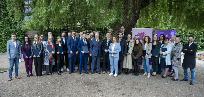 Grupa osób w różnym wieku, ubranych w eleganckie stroje, stoi na zewnątrz przed bujnym drzewem.
