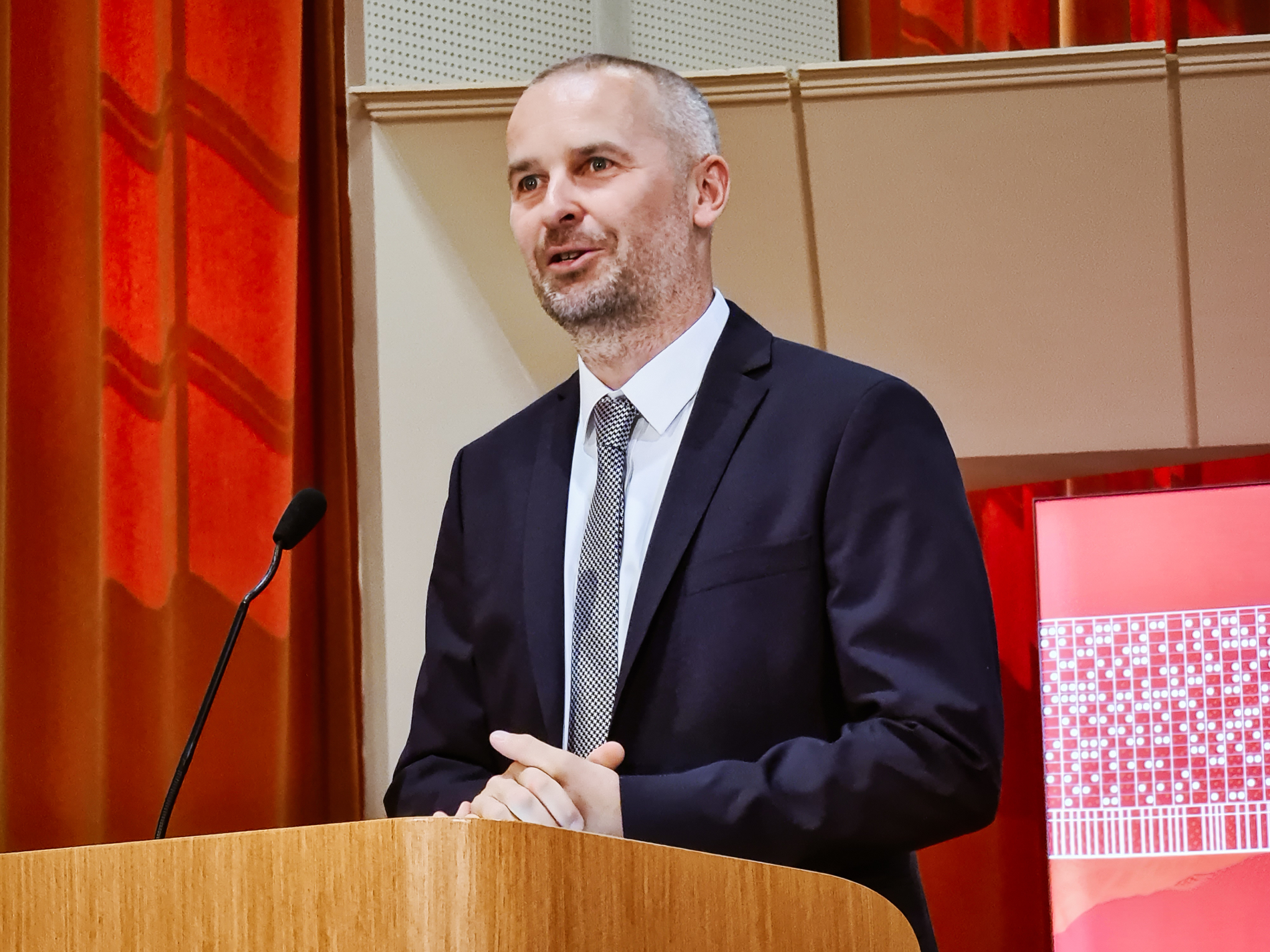 Mężczyzna w garniturze i krawacie stoi za drewnianą mównicą, przemawiając do publiczności. Po jego lewej stronie znajduje się mikrofon. 