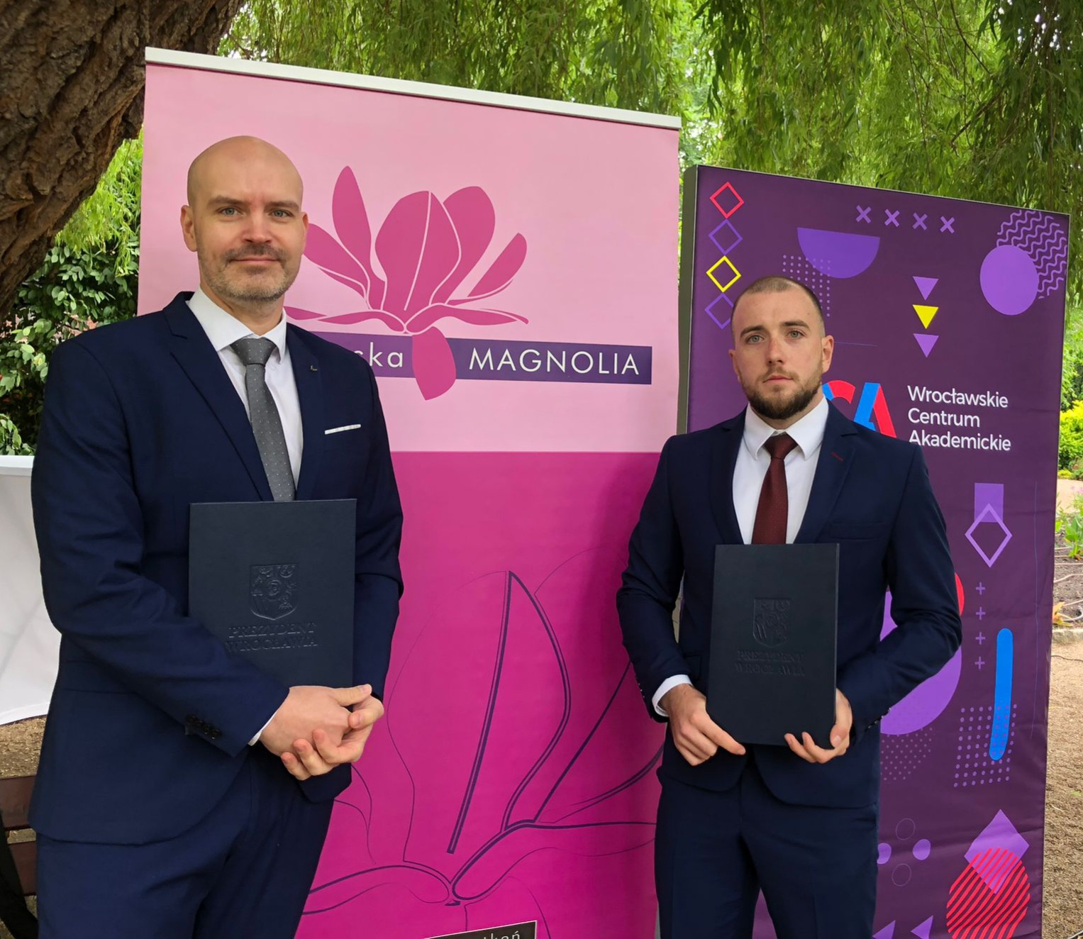 Stojący obok siebie dwaj mężczyźni w garniturach, trzymający teczki z dyplomami. W tle banery z logo „Magnolia” i „Wrocławskie Centrum Akademickie”.