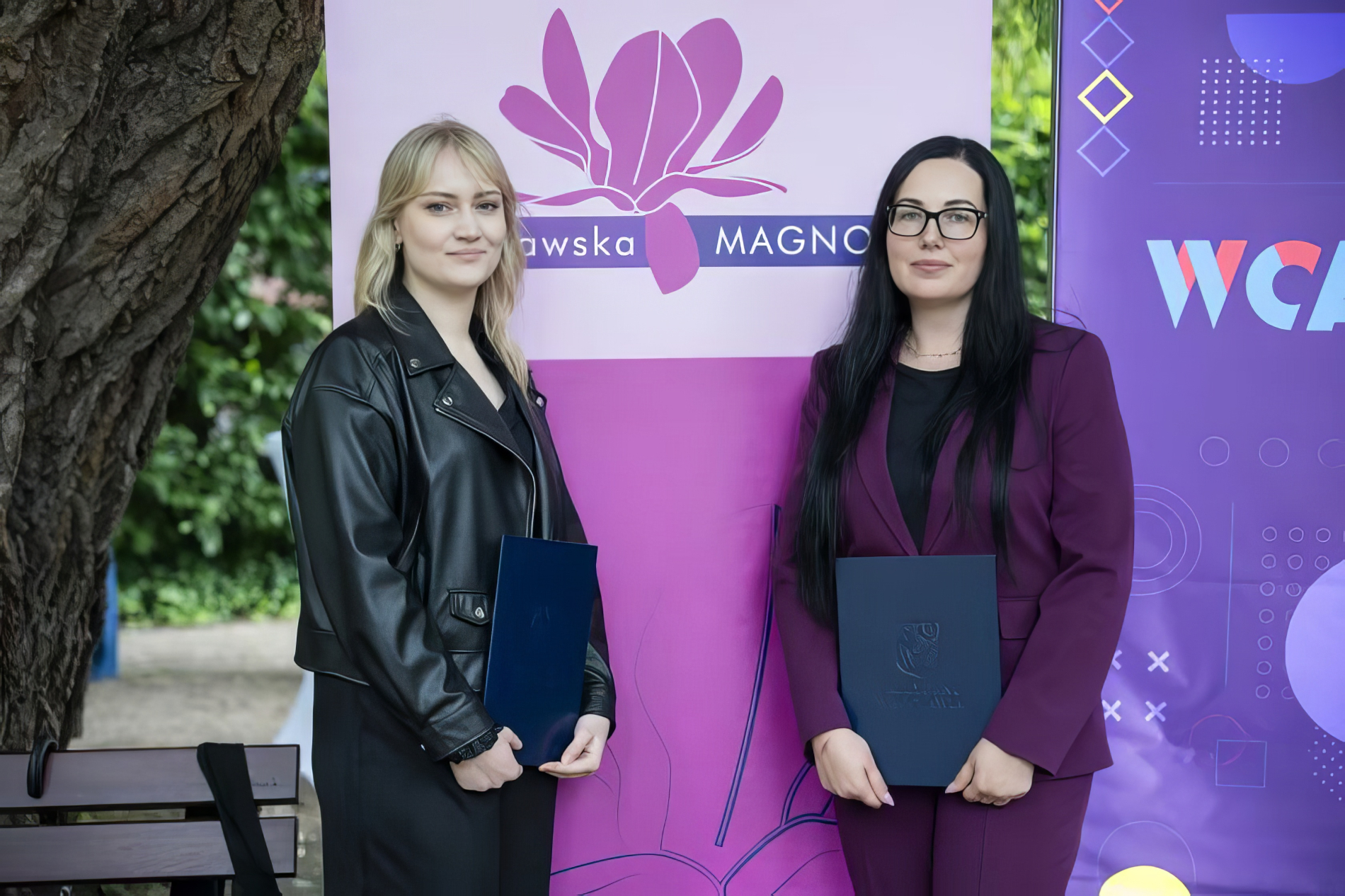 Dwie kobiety, ubrane w eleganckie stroje, stoją przed fioletowym banerem z napisem "Wrocławska Magnolia".