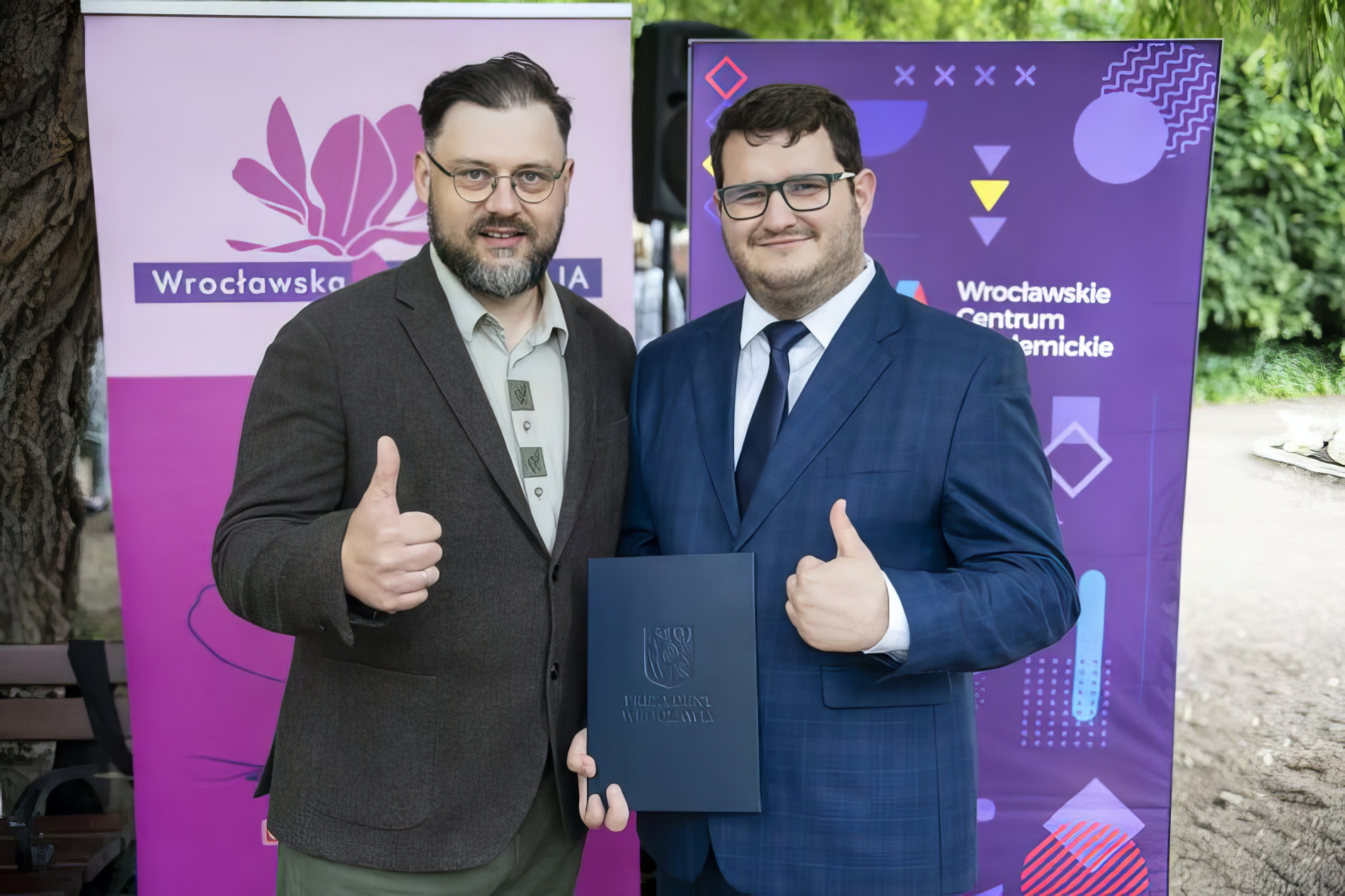 Mężczyzna z brodą i okularami w brązowej marynarce i koszuli stoi obok uśmiechniętego mężczyzny w garniturze i krawacie, również w okularach, trzymającego niebieską teczkę. Obaj pokazują kciuki w górę. 