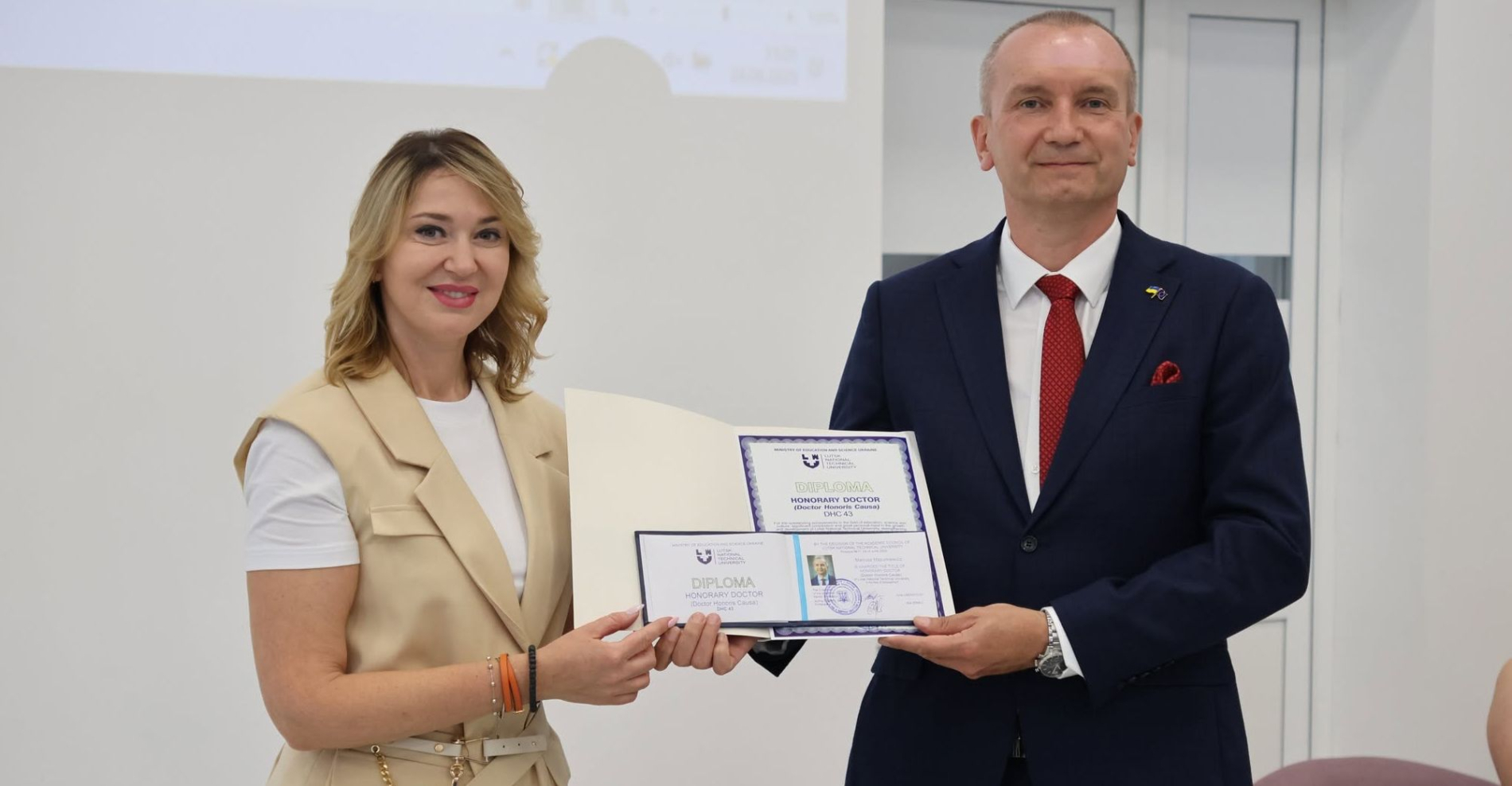 Pani dziekan wręcza dyplom honorowego doktora. Dyplom jest w białej teczce. Pani ubrana jest w jasną kamizelkę, pod którą ma biały T-shirt, a jej włosy są związane. Mężczyzna, który odbiera dyplom, ubrany jest w garnitur, białą koszulę i czerwony krawat.