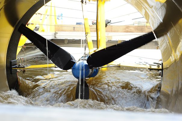 Widok z bliska na wirnik turbiny hydrokinetycznej w ruchu. Dwie czarne, szerokie łopaty obracają się, wzbijając brudną wodę rzeki.