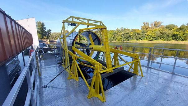 Duża, żółta turbina wodna, zamontowana na metalowym pokładzie barki pływającej po rzece, otoczona zieloną roślinnością.