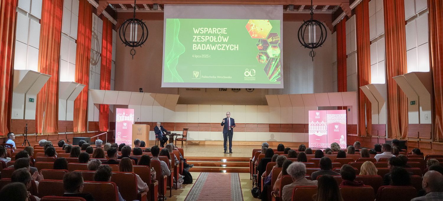 Zdjęcie przedstawia dużą aulę Uniwersytetu Wrocławskiego podczas konferencji "Wsparcie Zespołów Badawczych". Na scenie przemawia mężczyzna w garniturze. Przed nim siedzi publiczność. Na ekranie za prelegentem widoczna jest prezentacja z logo uczelni i datą "4 lipca 2025 r.". W tle charakterystyczne wnętrze auli z wysokimi oknami.