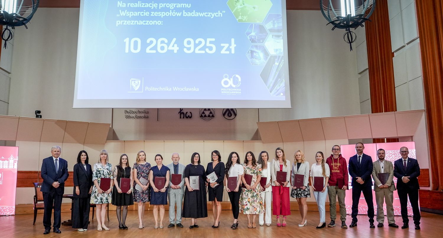 Zdjęcie grupowe przedstawia około 20 osób stojących w rzędzie na scenie. Wszyscy trzymają dokumenty lub teczki. W tle widoczny jest duży ekran z napisem "Na realizację programu „Wsparcie zespołów badawczych” przeznaczono: 10 264 925 zł" oraz logo Politechniki Wrocławskiej.