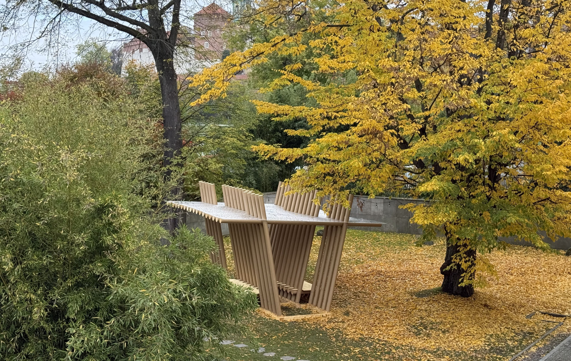 Zdjęcie pawilonu z rur papierowych stojącego wśród drzew, w tle zamek na Wawelu.