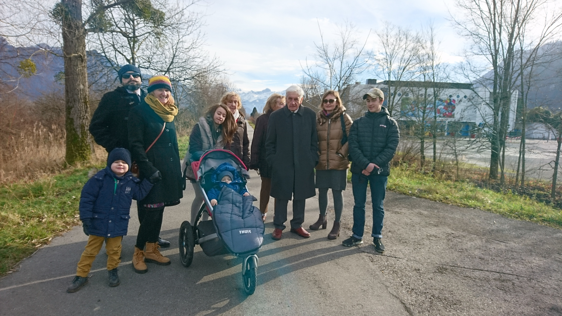 Duże, rodzinne zdjęcie. Przedstawia trzech dorosłych mężczyzn, cztery dorosłe kobiety i dwójkę małych dzieci w wózku i obok wózka. Wszyscy patrzą w stronę obiektywu, są ubrani w kurtki, czapki, szaliki. W tle widać nagie drzewa, budynki i góry.