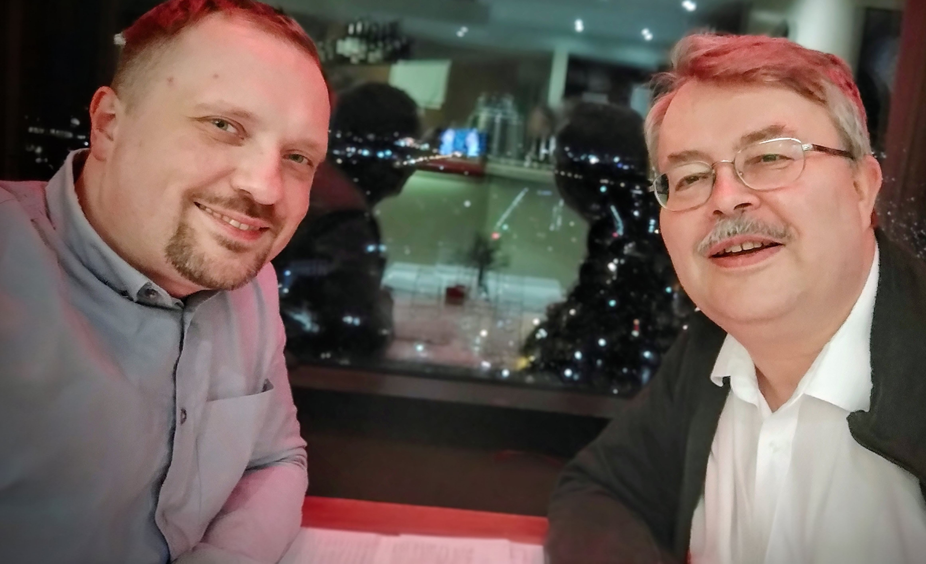 Dwóch mężczyzn siedzących przy stole, w tle widoczna panorama nocnego miasta za oknem.