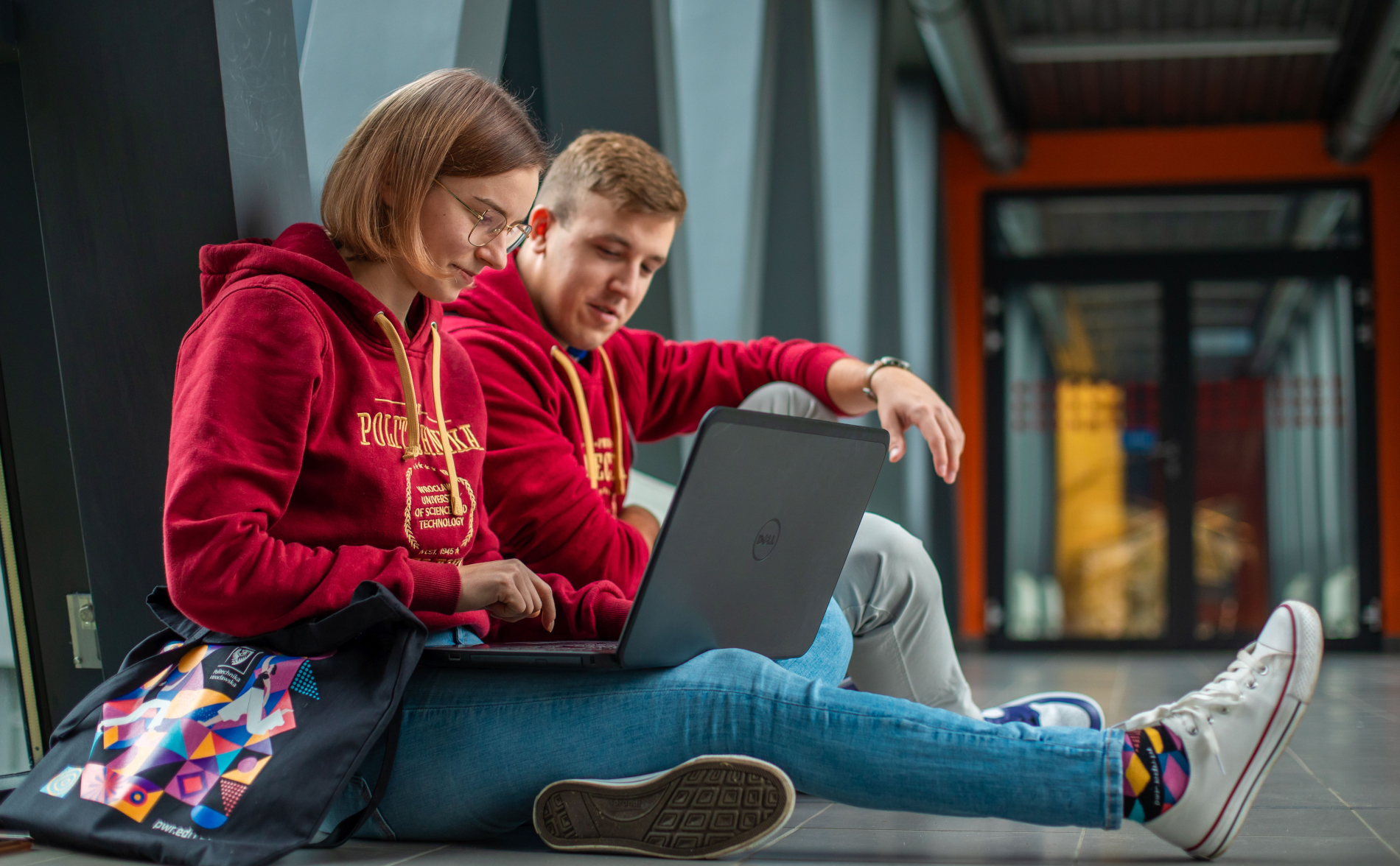 Dwoje młodych ludzi, studentka w okularach i student, siedzą na podłodze korytarza i pochylają się nad laptopem. Oboje mają na sobie czerwone bluzy z logo uczelni. W tle widać nowoczesne wnętrze budynku uniwersyteckiego.