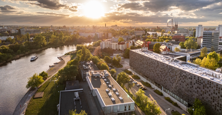 Panoramiczny widok na Wrocław o zachodzie słońca, z rzeką Odrą po lewej stronie i nowoczesnymi budynkami oraz zielenią rozciągającą się w kierunku horyzontu. Słońce, nisko na niebie, tworzy długie cienie i refleksy na wodzie.