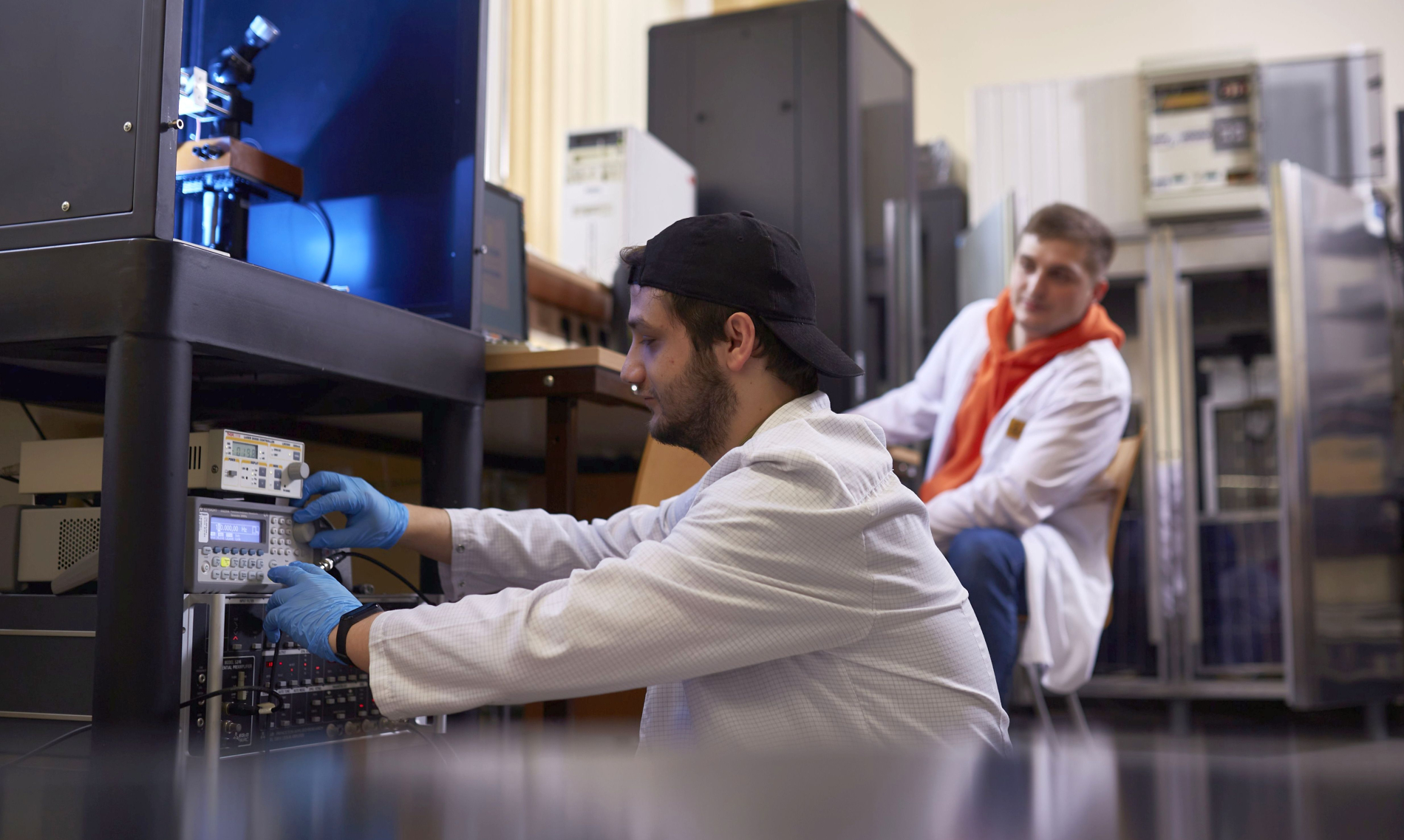 Mężczyzna w fartuchu laboratoryjnym i rękawiczkach reguluje aparaturę pomiarową, a drugi siedzi w tle w laboratorium naukowym.