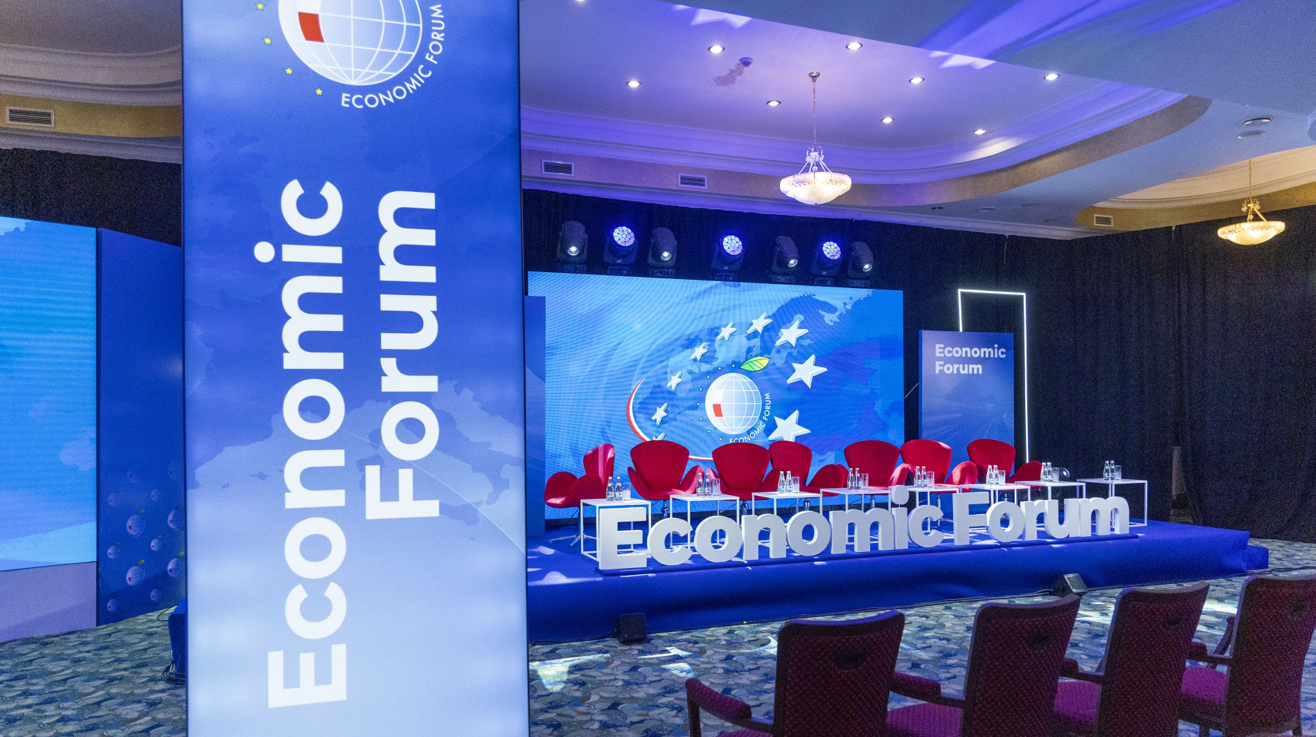 Pusta scena przygotowana na Forum Ekonomiczne w Karpaczu. W centrum znajdują się czerwone fotele dla prelegentów ustawione w rzędzie, z przodu stoły z butelkami wody. Po bokach stoją duże niebieskie ekrany z napisem "Economic Forum".