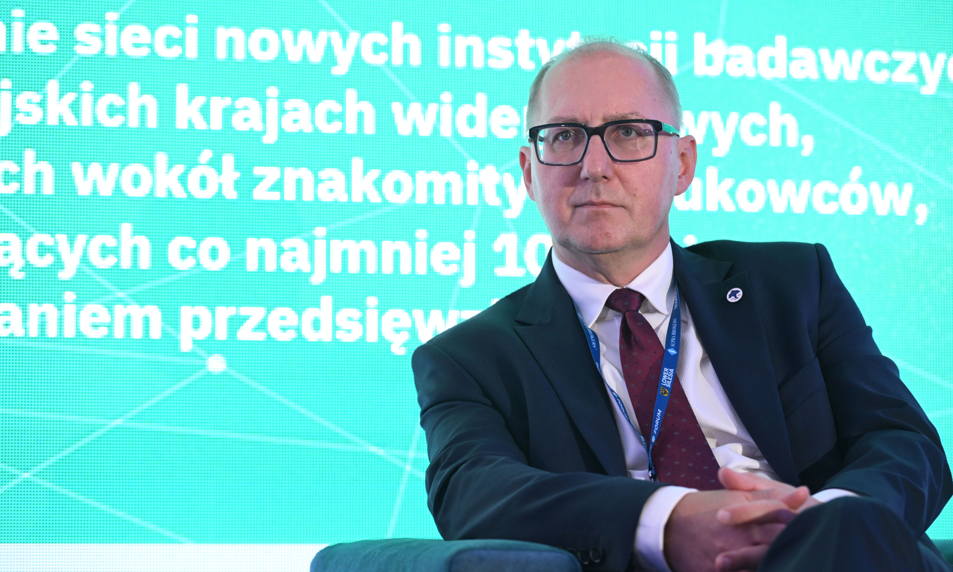 Mężczyzna w okularach, marynarce i krawacie siedzi na krześle, patrząc w prawo. Na tle widać fragment zielonego ekranu z niewyraźnym tekstem w języku polskim.